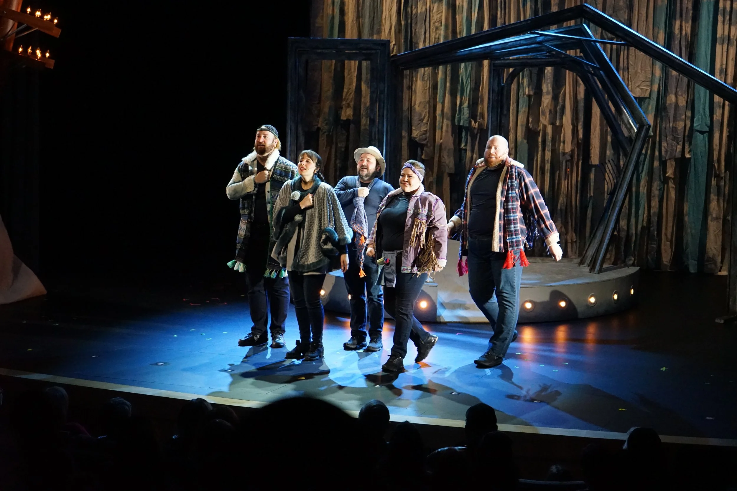 Groupe de six acteurs sur scène lors d'une représentation théâtrale, vêtus de costumes décontractés et colorés, dans une mise en scène inspirée de la nature.