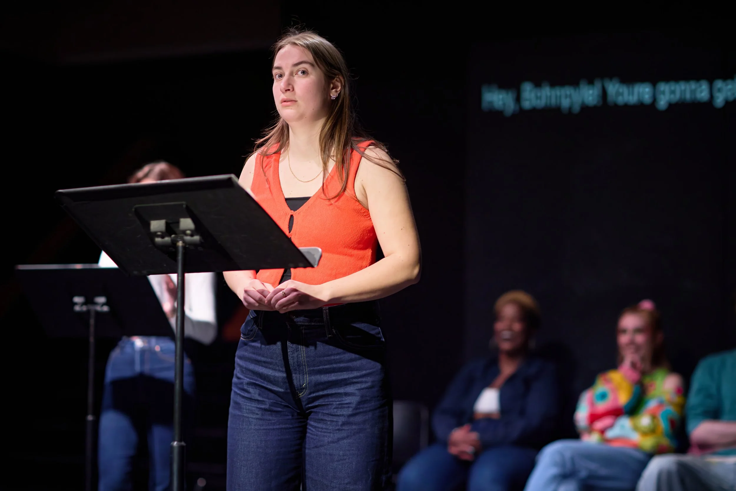 Jeune femme debout devant un pupitre, portant un haut orange, lors d'un spectacle ou d'une présentation avec un groupe en arrière-plan.