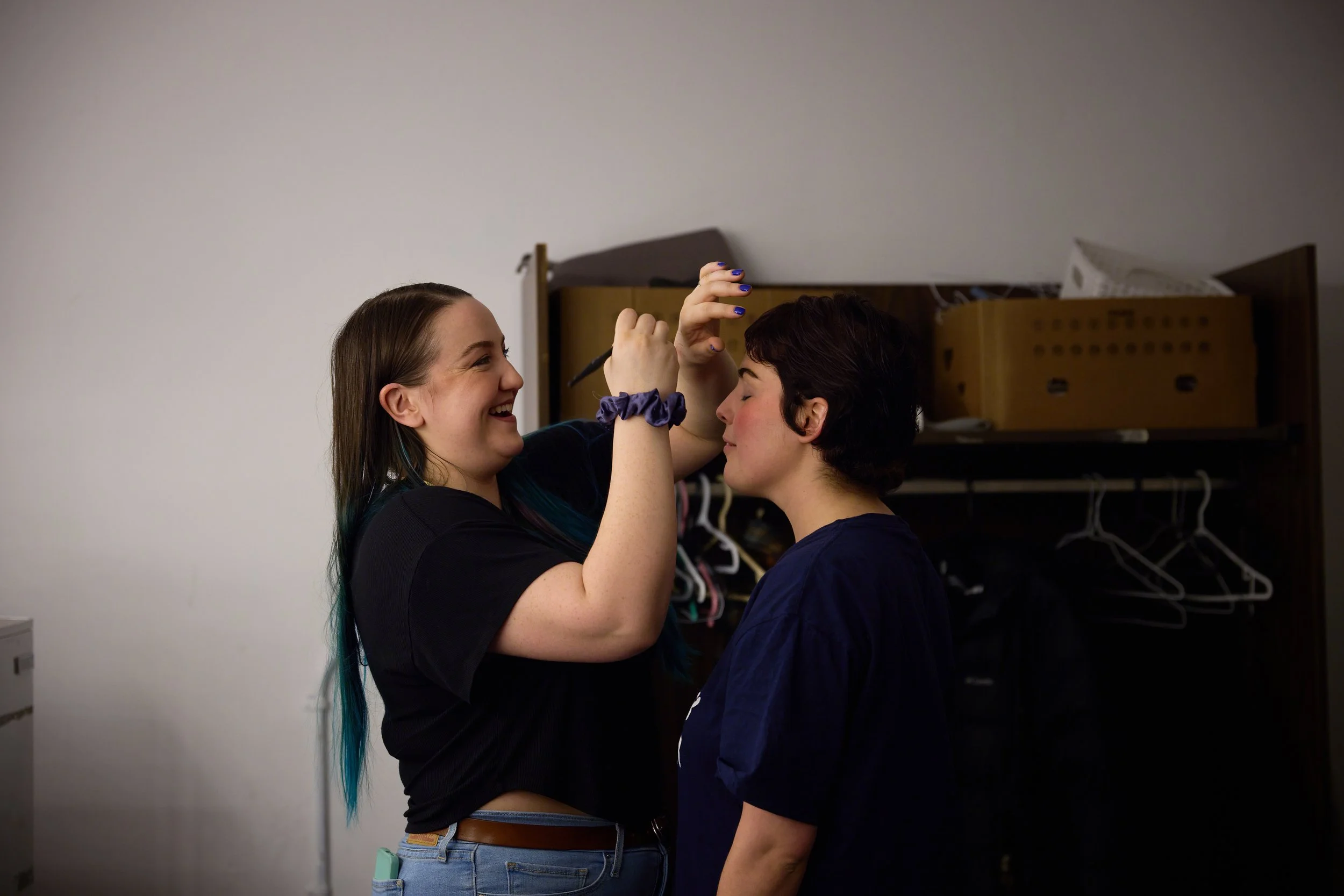 Deux jeunes femmes, l'une souriante et l'autre ayant les yeux fermés, sont en train de se maquiller ou de se préparer devant une armoire de rangement dans une pièce. La femme à gauche utilise un pinceau ou un maquillage sur le visage de la femme à dr