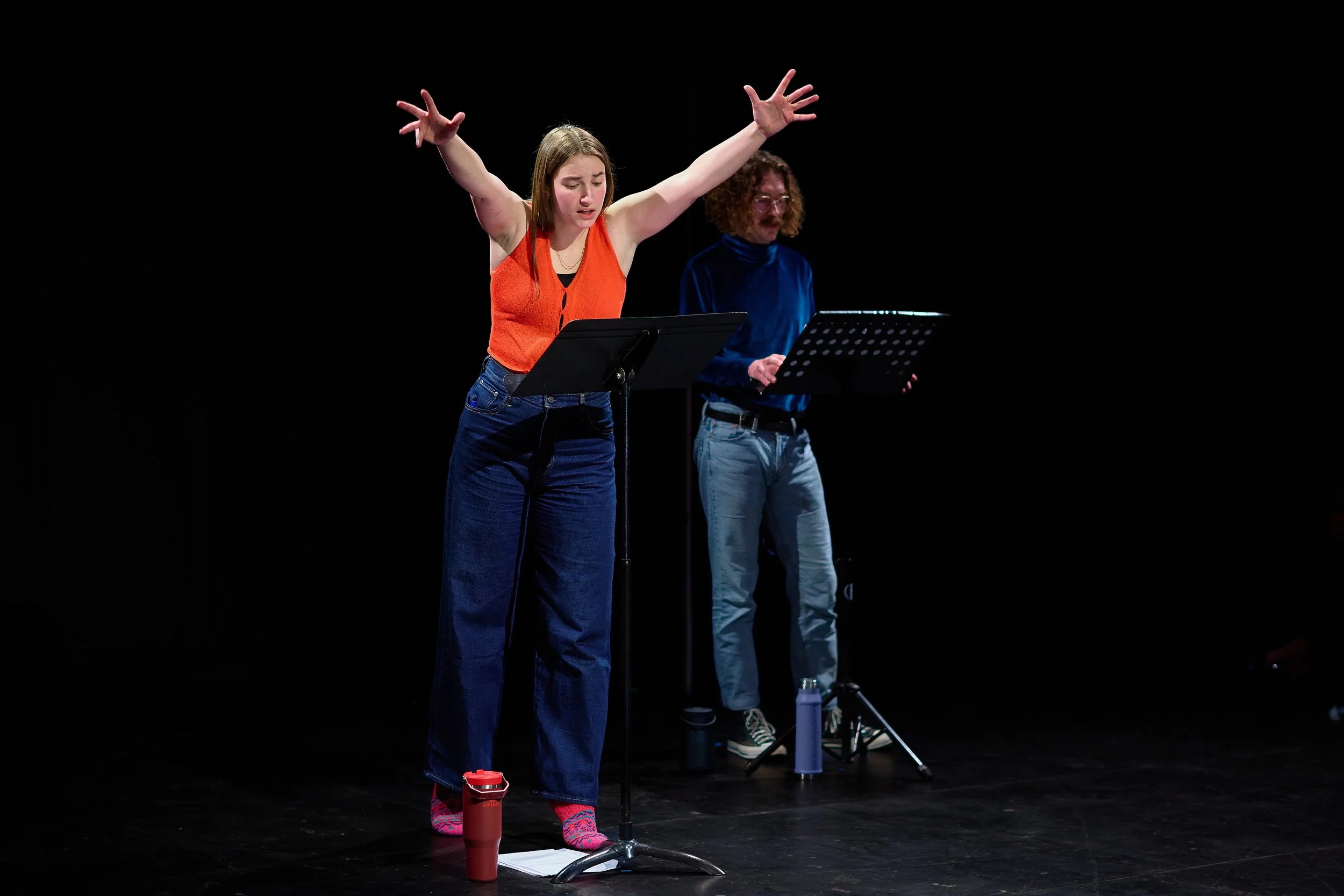 Une jeune femme avec un débardeur orange et un pantalon large bleu, les bras levés, semble en pleine performance ou rehearsal sur scène, avec un homme en arrière-plan jouant du piano.