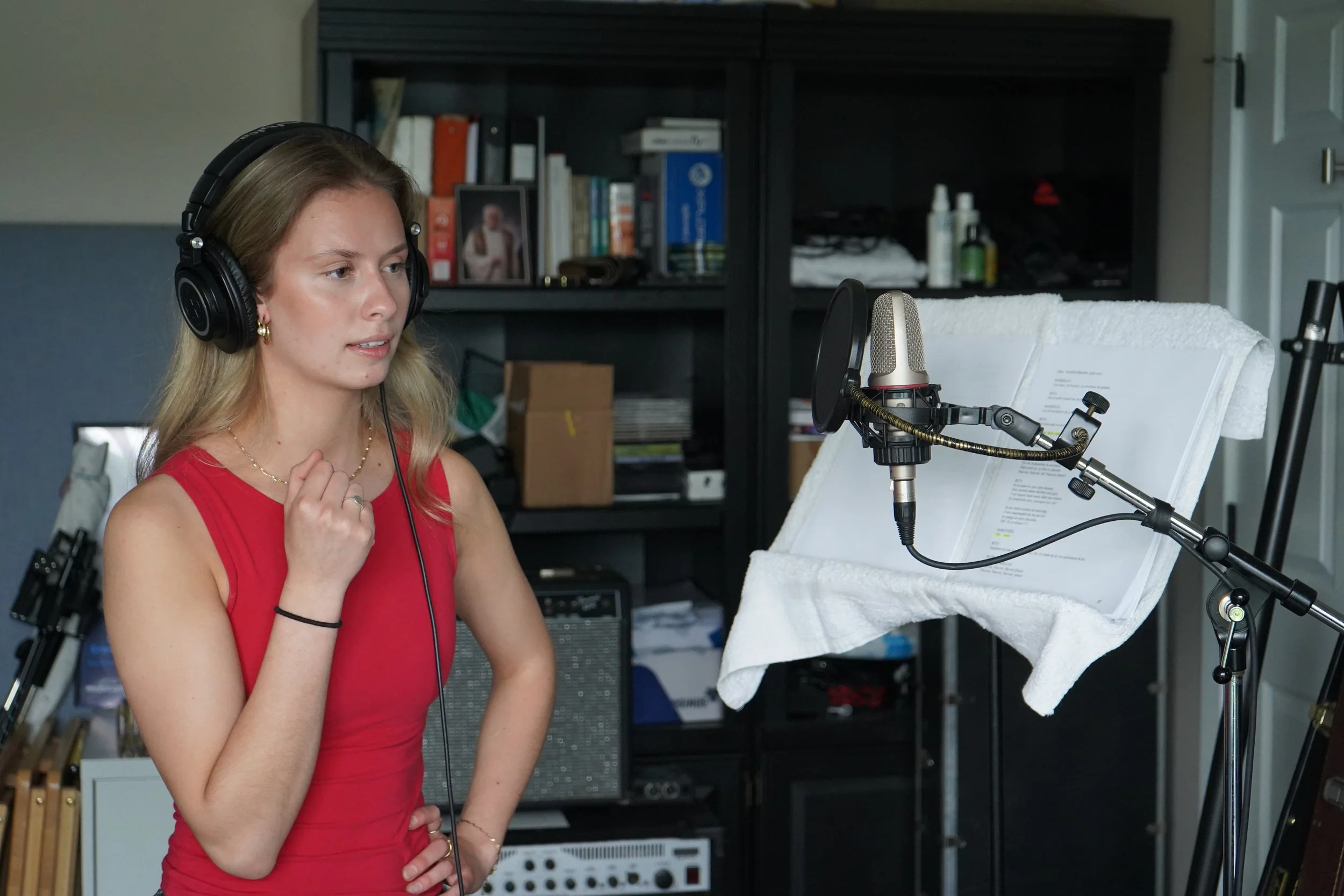 Une jeune femme portant un haut rouge, des écouteurs et une chaîne en or, se tient devant un microphone dans un studio d'enregistrement, avec une partition sur un support. En arrière-plan, une étagère noire avec des livres, des boîtes, des produits d