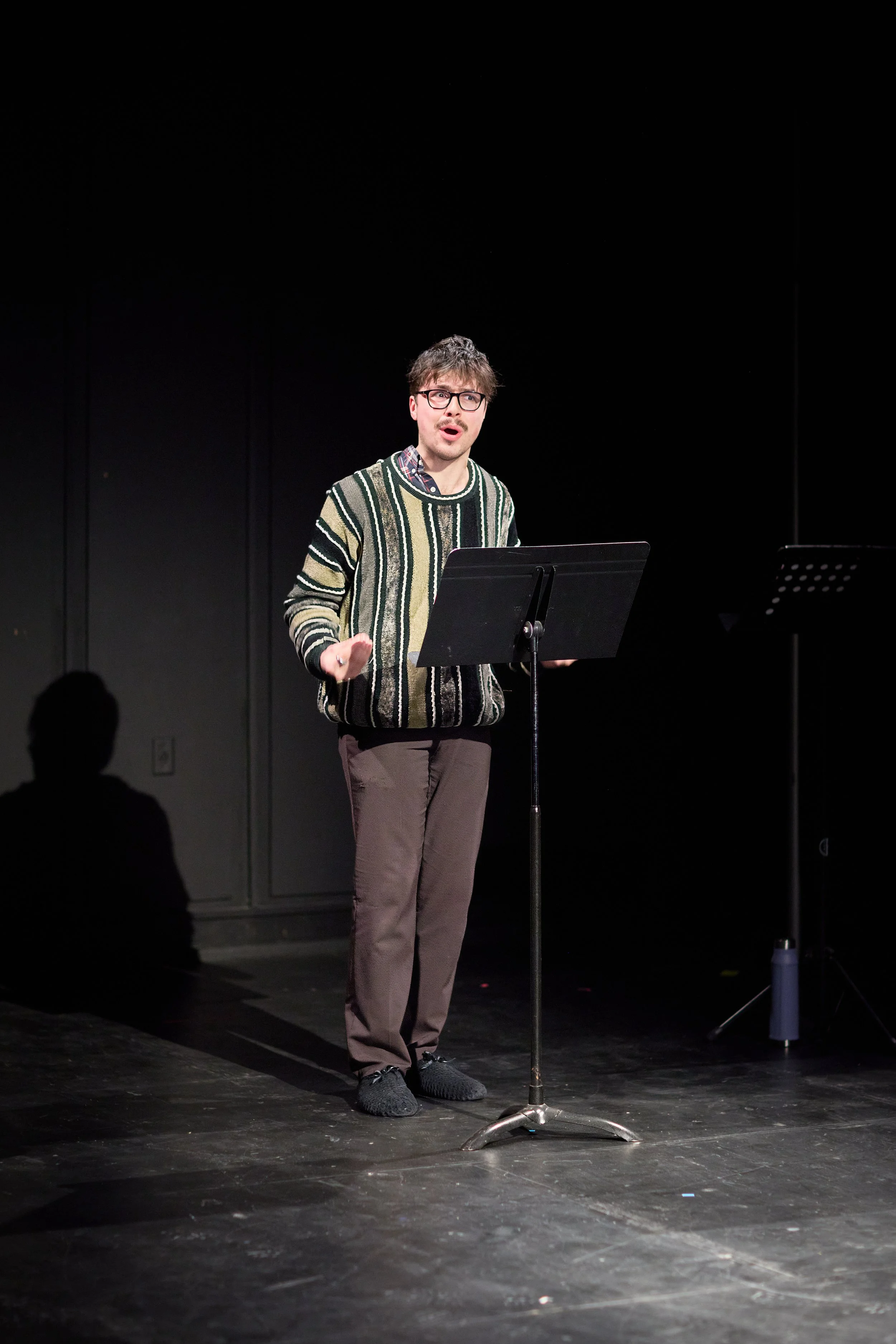 Un homme debout devant un pupitre noir sur scène, parlant ou chantant, dans un endroit sombre avec un fond noir.