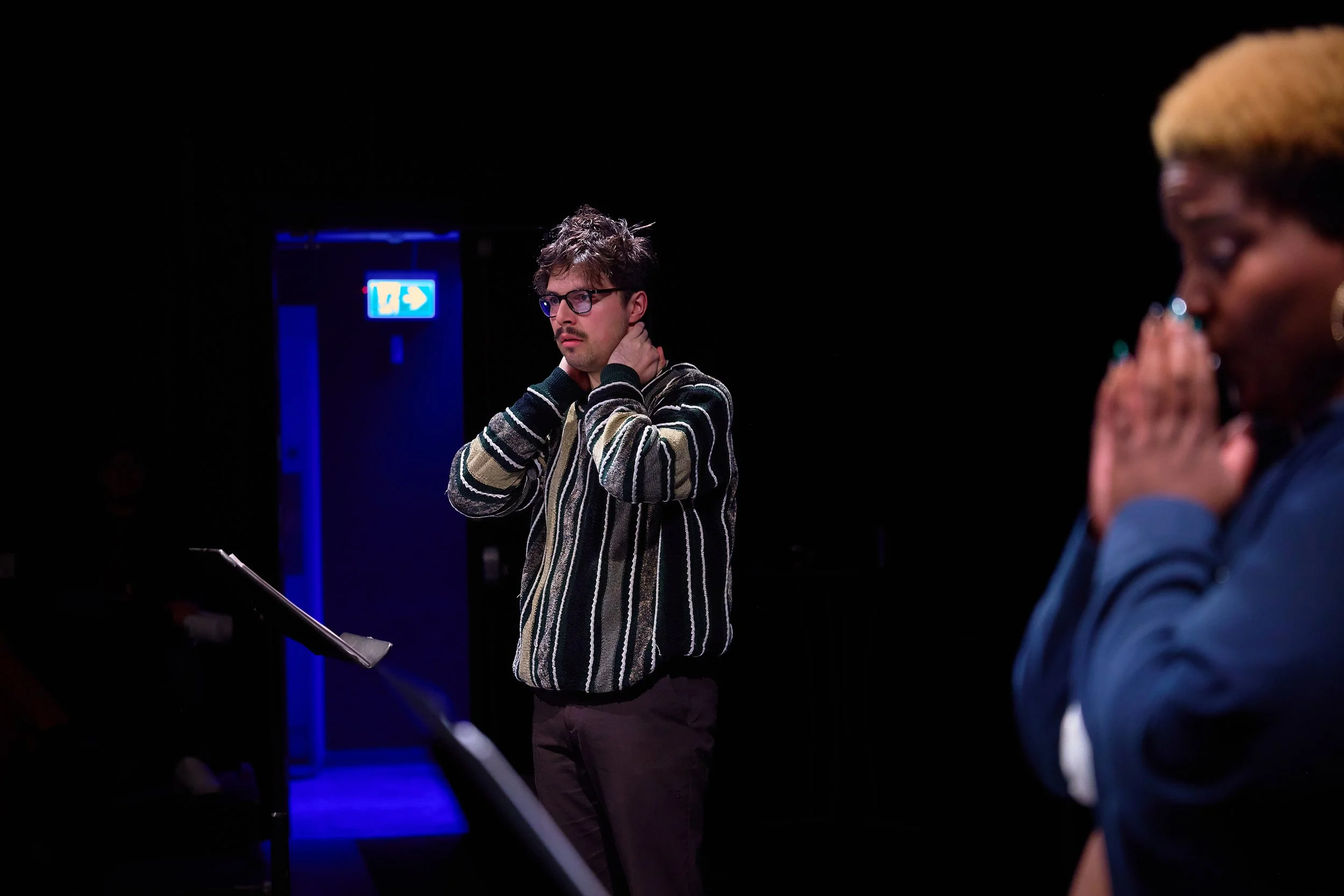 Deux personnes se tiennent dans une pièce sombre, la personne à gauche est un homme avec des lunettes, portant un pull rayé, semblant réfléchir ou préoccupé, la personne à droite, une femme aux cheveux courts, portant une veste bleue, a les mains joi