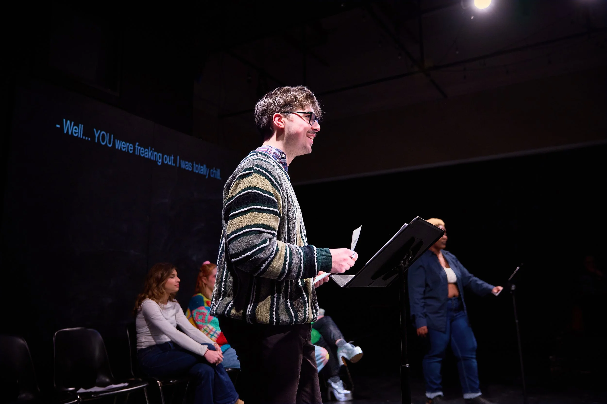 Un homme avec des lunettes, debout devant un pupitre dans une salle sombre, parlant ou lisant avec un groupe de personnes assises derrière lui. L'arrière-plan montre un écran avec un sous-titre en anglais.