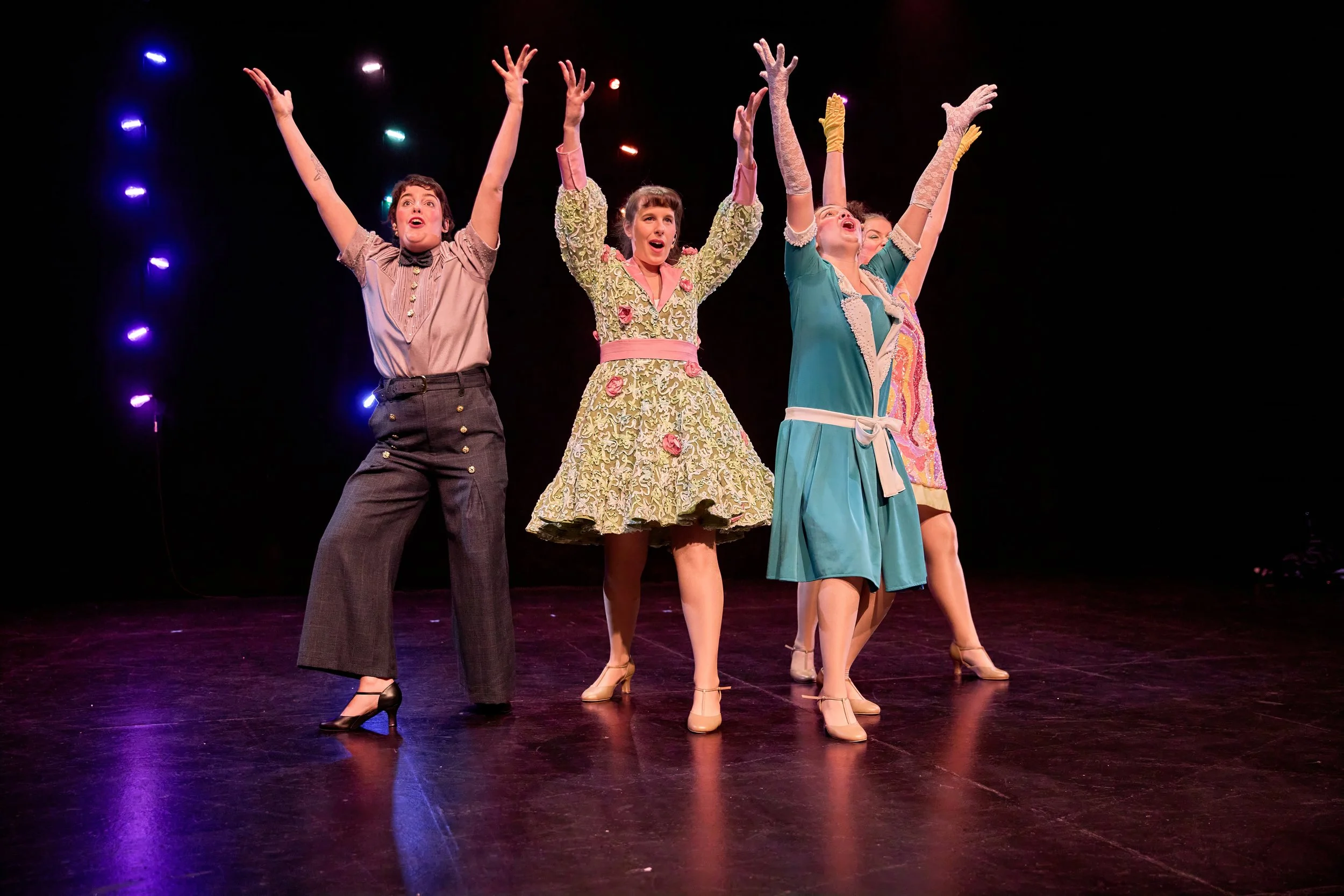 Cinq femmes en costume de théâtre chantent et dansent sur scène avec des bras levés dans un spectacle.