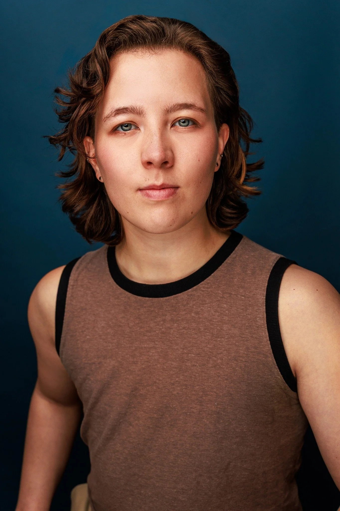 Jeune femme aux cheveux bruns bouclés, portant un débardeur marron avec des bandes noires, pose contre un fond bleu.