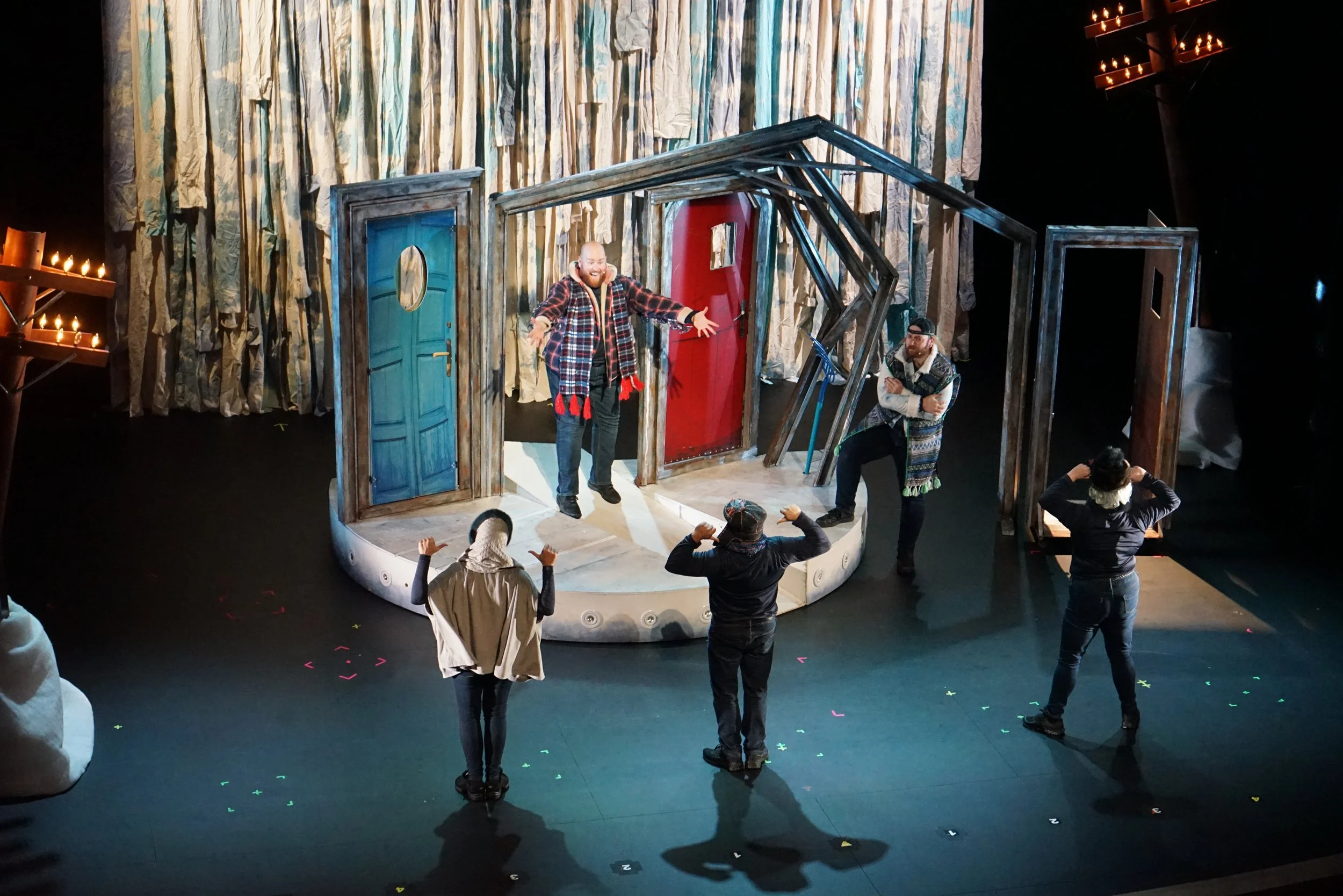 Scène de théâtre avec deux acteurs sur scène et trois personnes en répétition ou en séance d'entraînement en face, dans une pièce décorée de portes et d'une structure en bois. Le décor semble évoquer une maison ou un intérieur.