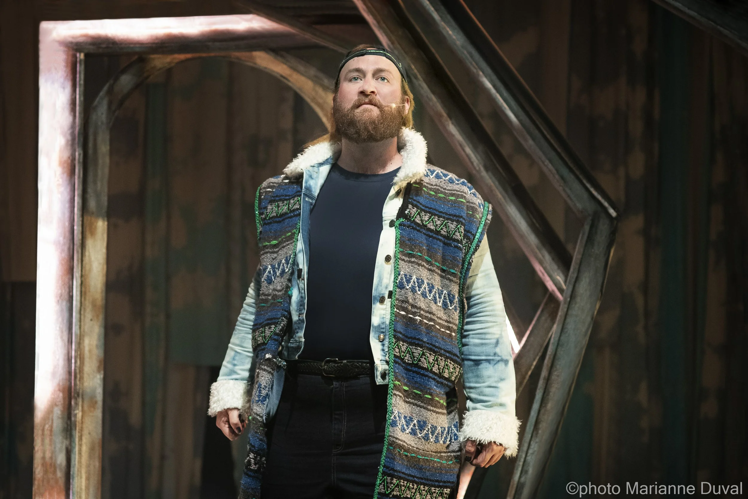 Un homme avec une barbe portant un manteau en patchwork coloré, une casquette, et un micro près de la bouche, se tenant devant une structure métallique dans un environnement sombre.