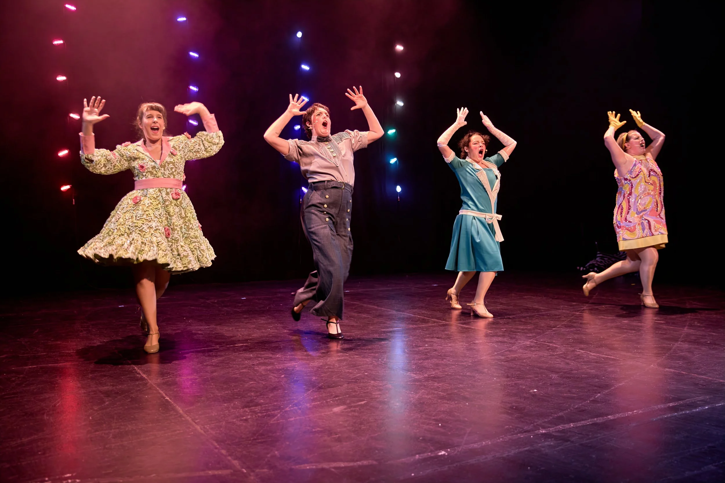Quatre femmes en costume vintage dansant sur scène avec des lumières colorées en arrière-plan.