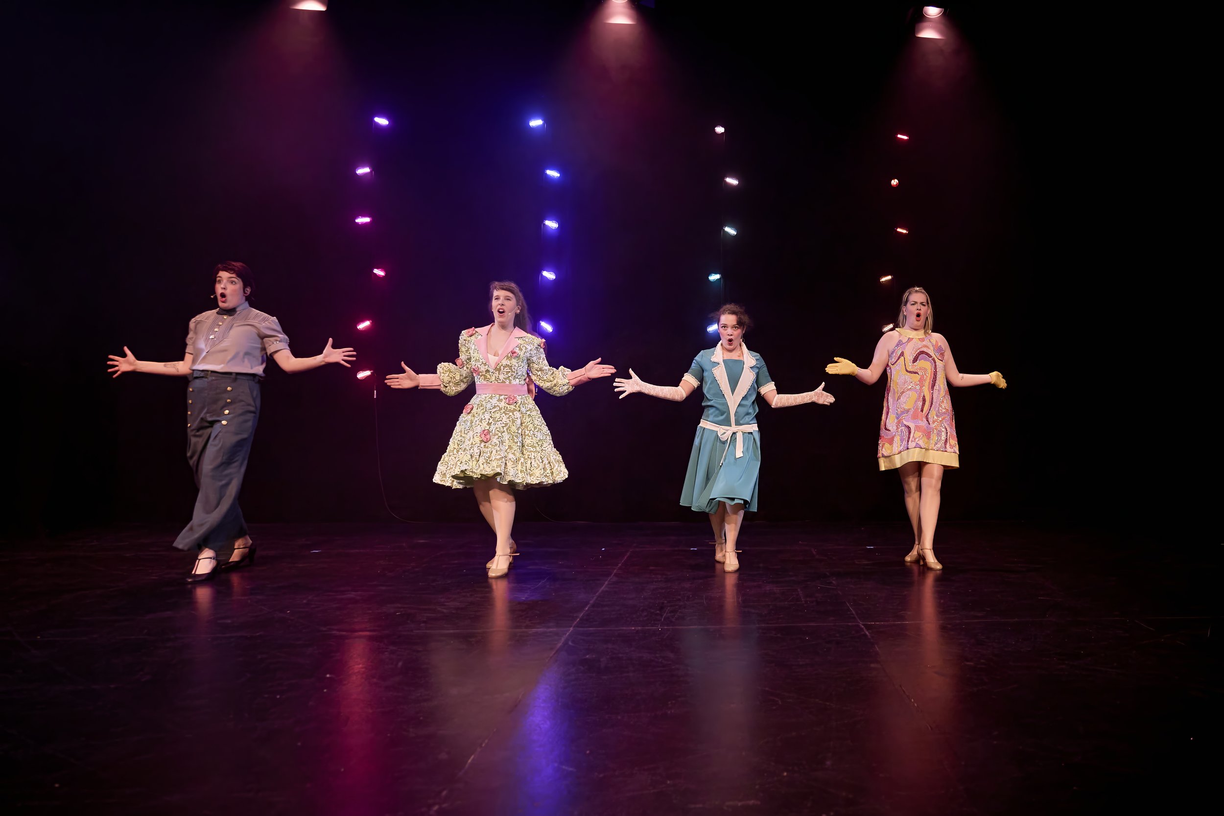 Quatre femmes en costume de scène chantent sur une scène sombre avec un fond éclairé de lumières colorées.