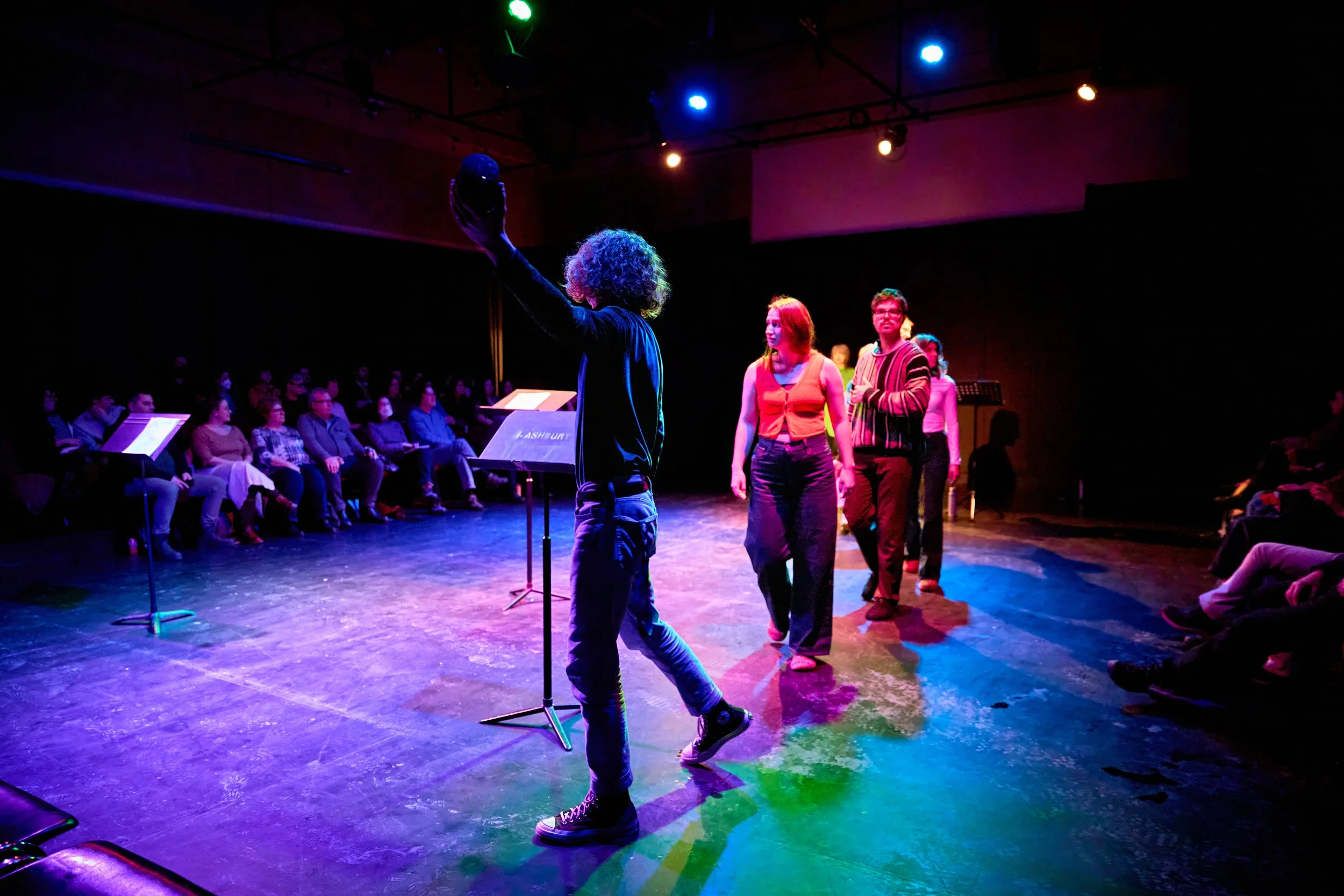 Une scène de théâtre avec un groupe de personnes en costume, colorées et éclairées par des lumières artistiques, regardant un spectacle.