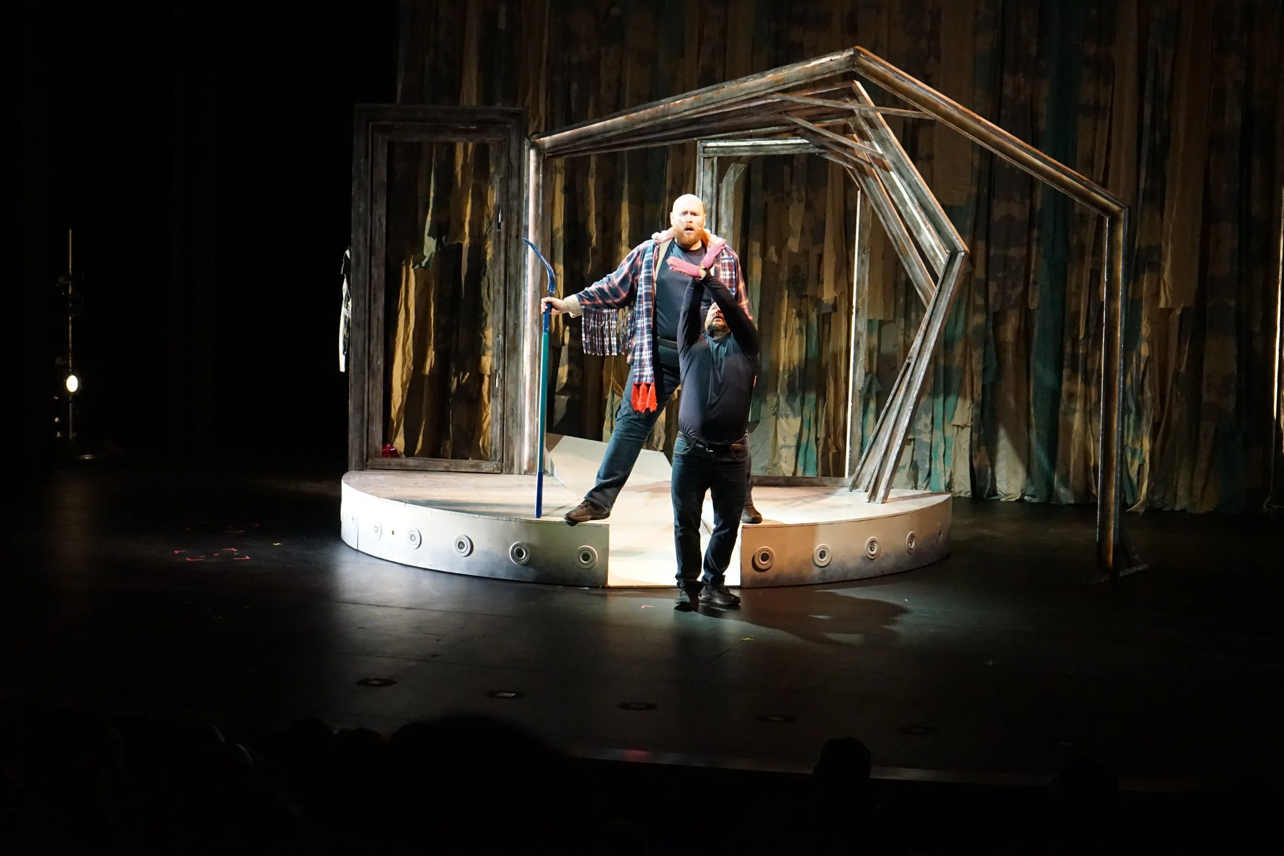 Deux hommes sur une scène de théâtre, l'un en geste expressif, l'autre tenant un bâton, dans un décor minimaliste avec des structures en bois et un fond en tissu coloré.