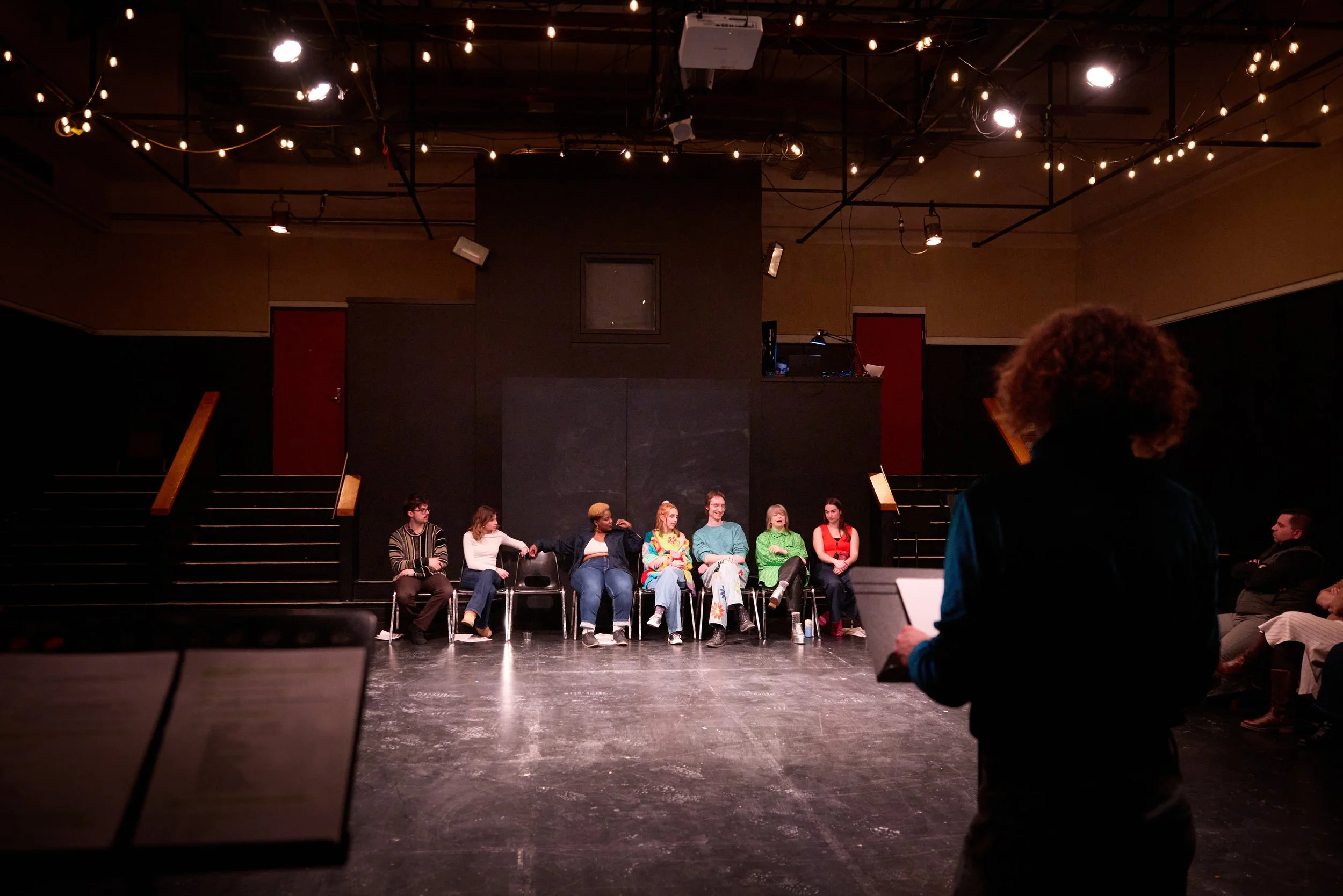 Groupe de personnes assises sur scène dans un théâtre ou studio, avec un éclairage chaud et décoration de guirlandes lumineuses, en attente ou en répétition.