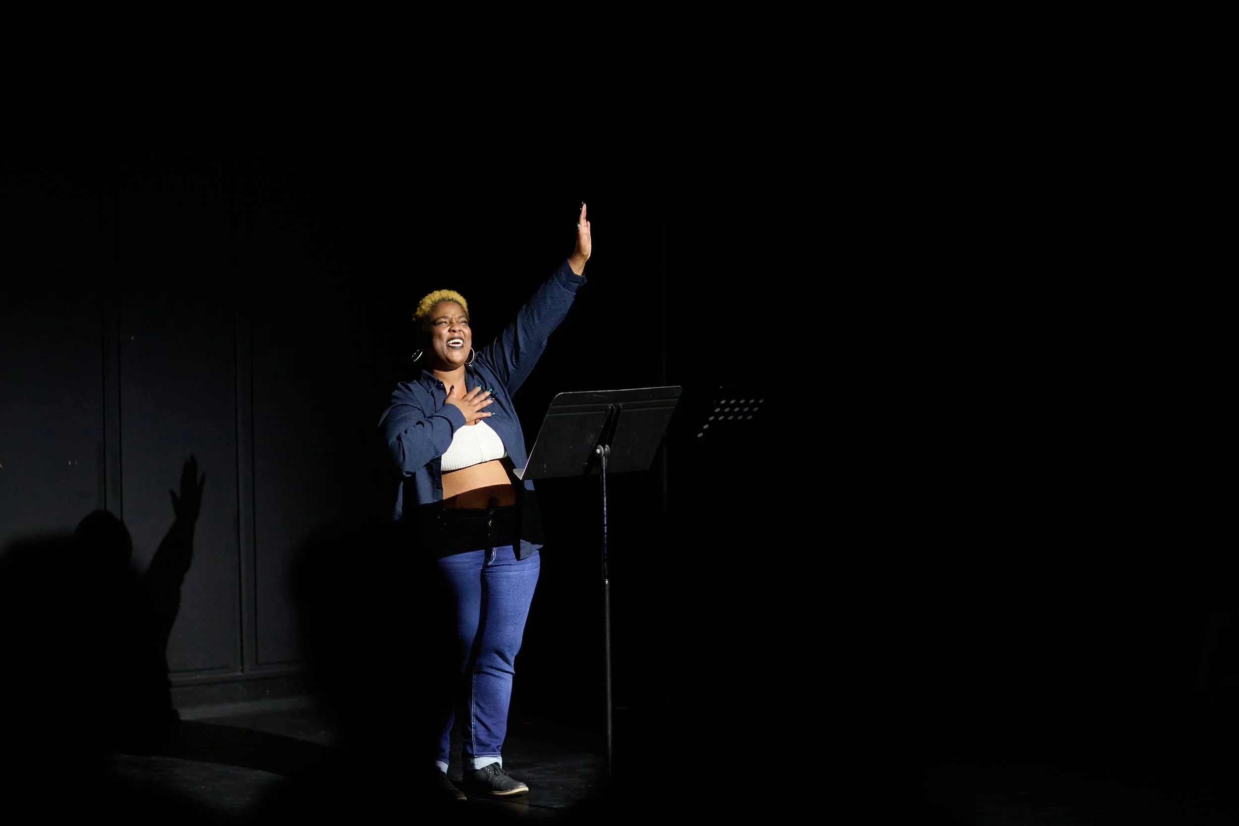 Une femme sur scène, jusqu'à la poitrine, en train de chanter ou de réciter, avec une grande émotion, dans un environnement sombre, éclairée par un spot lumineux.