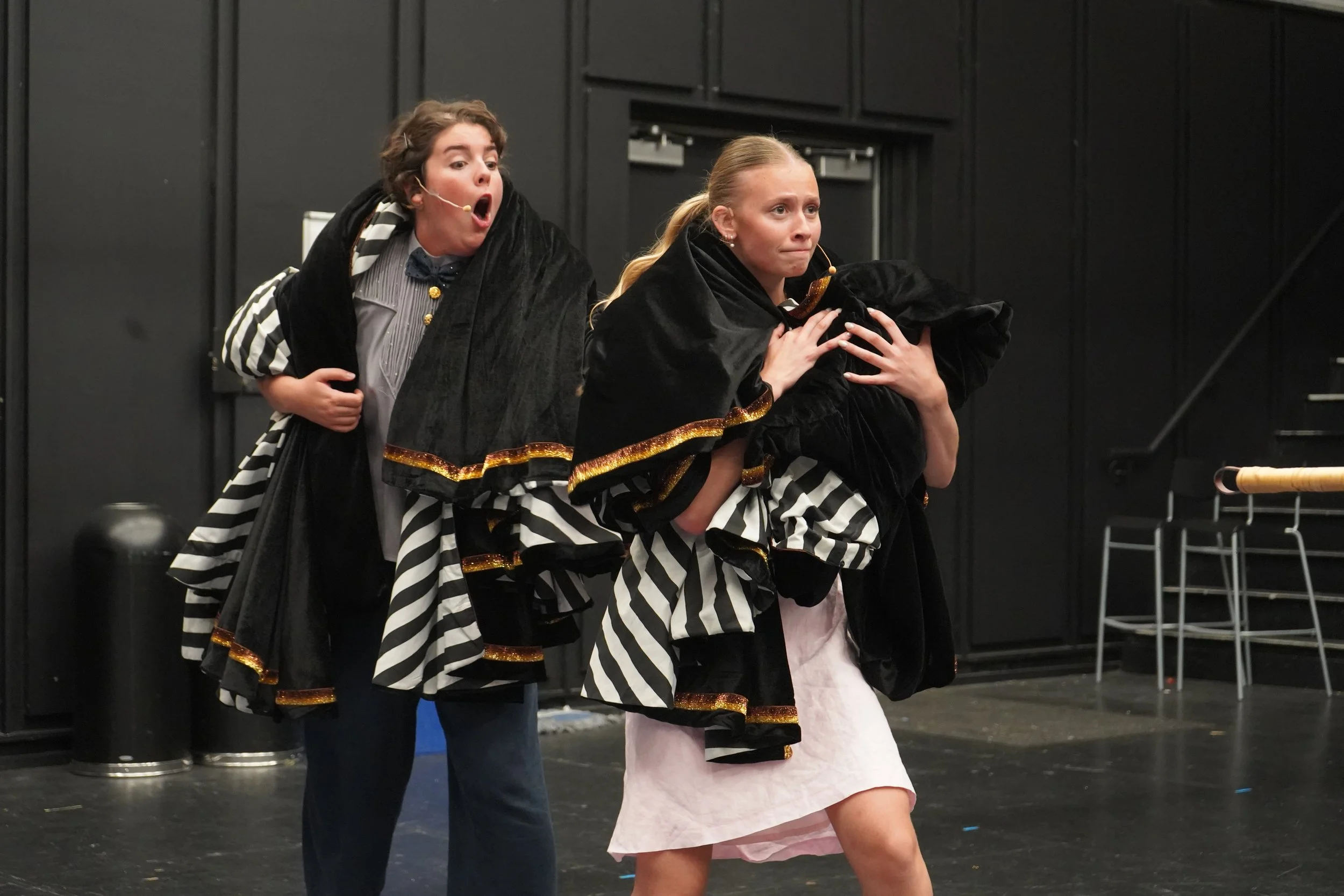 Deux femmes en costumes de théâtre expressifs, avec des robes à rayures noires et blanches et des capes noires avec des bordures dorées, sur scène lors d'une performance.