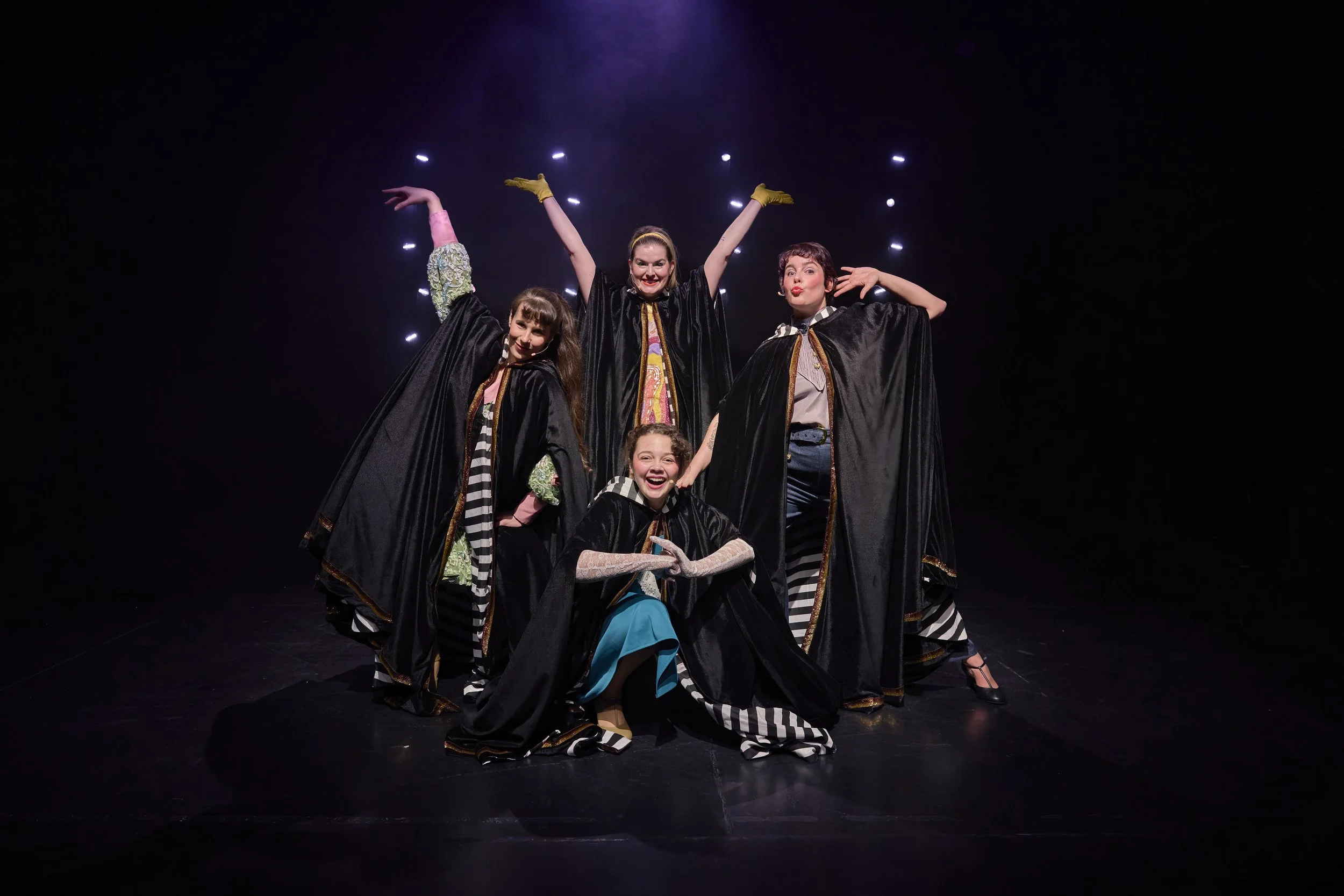 Créations In Vivo. Groupe de cinq jeunes filles dans une scène de spectacle, vêtues de robes noires avec des détails colorés et à rayures, posant théâtralement sous des lumières de scène