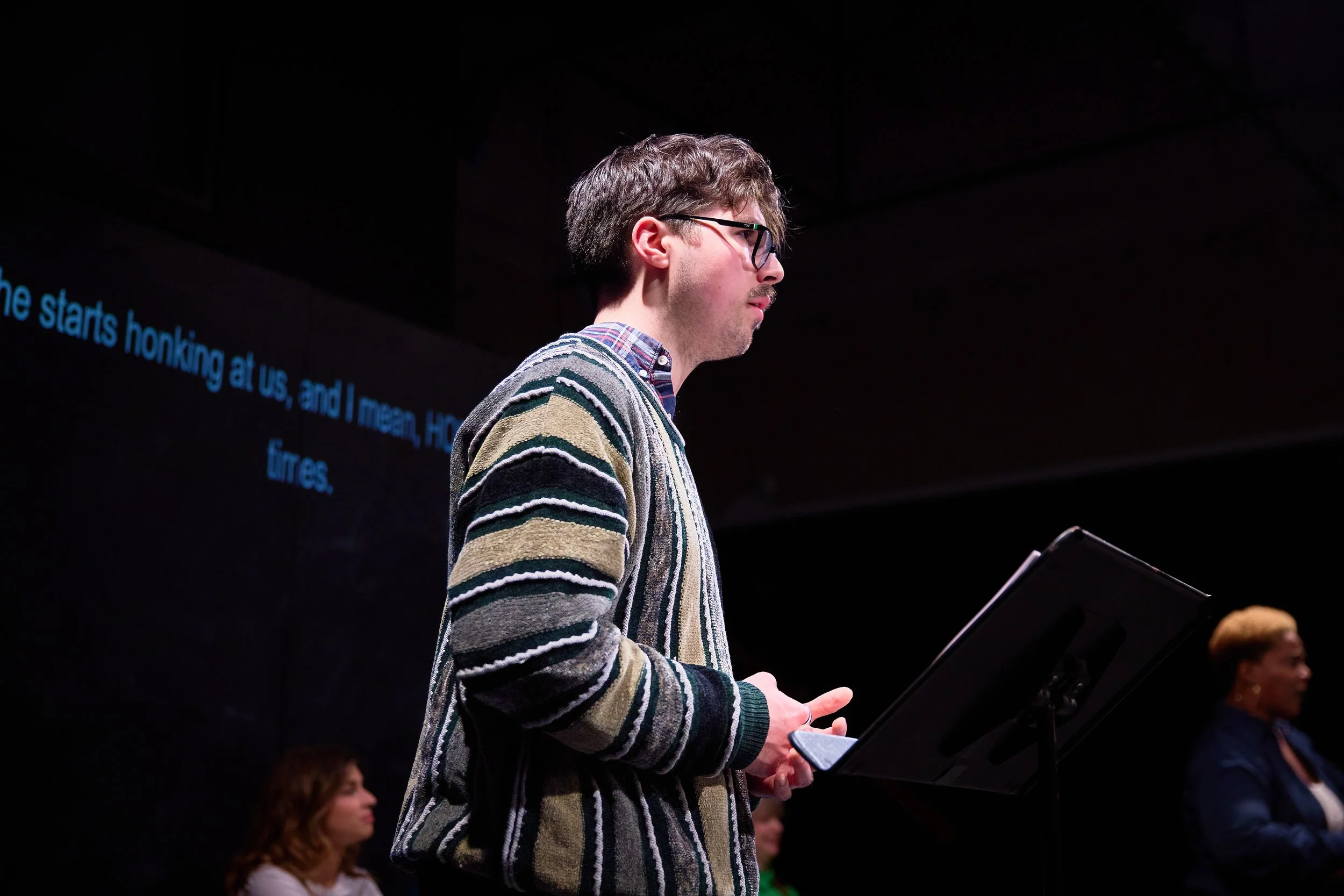 Jeune homme with glasses et tricot rayé, tenant un tablette, devant un écran noir avec texte en anglais en arrière-plan, probablement lors d'une présentation ou d'une conférence.