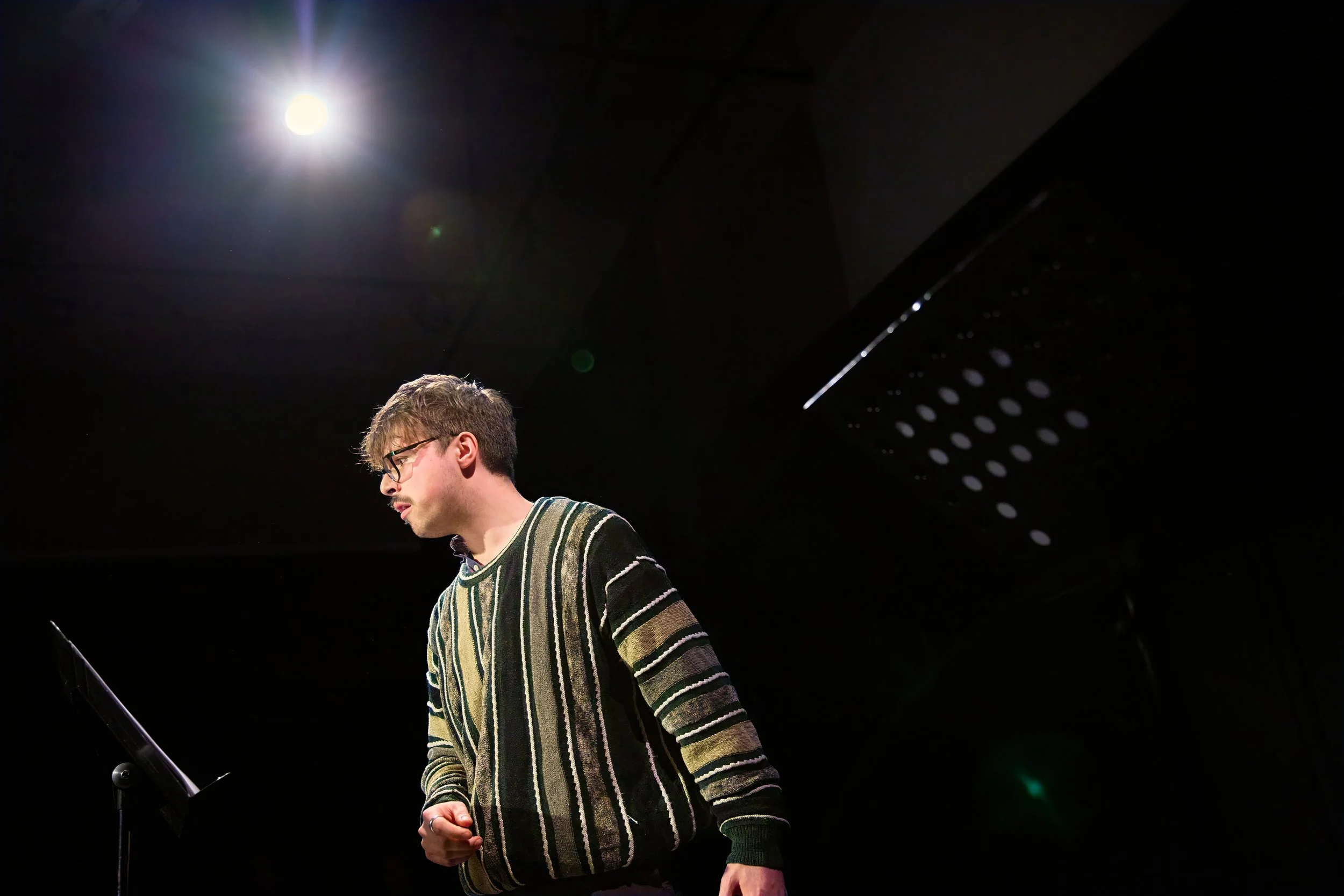 Jeune homme portant des lunettes et un pull rayé, se tenant à côté d'un pupitre sur scène, sous un seul projecteur.
