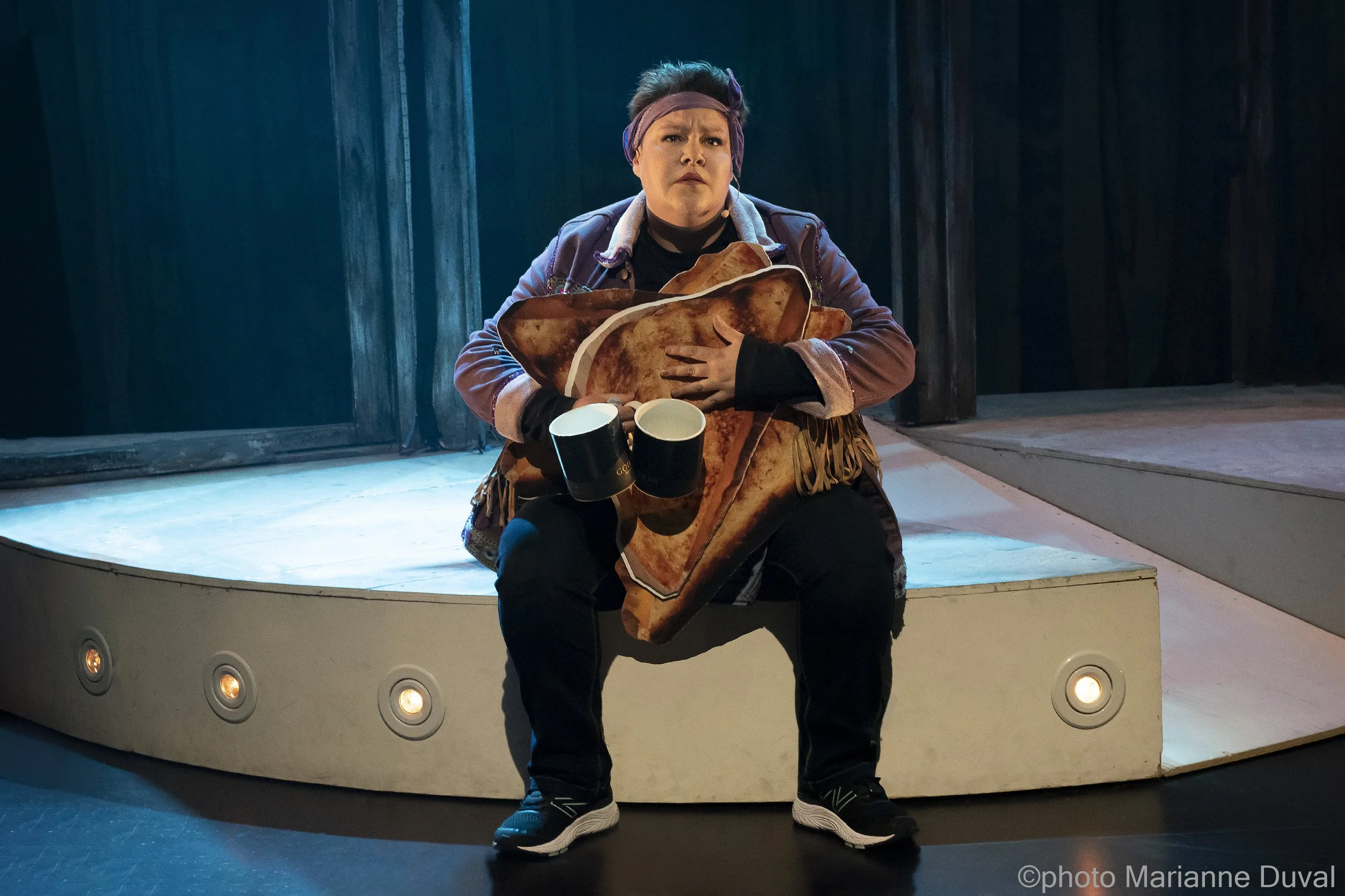 Une femme assise sur une scène, tenant un grand sac de chips et deux mugs, portant un bandeau violet sur la tête, dans un décor sombre.