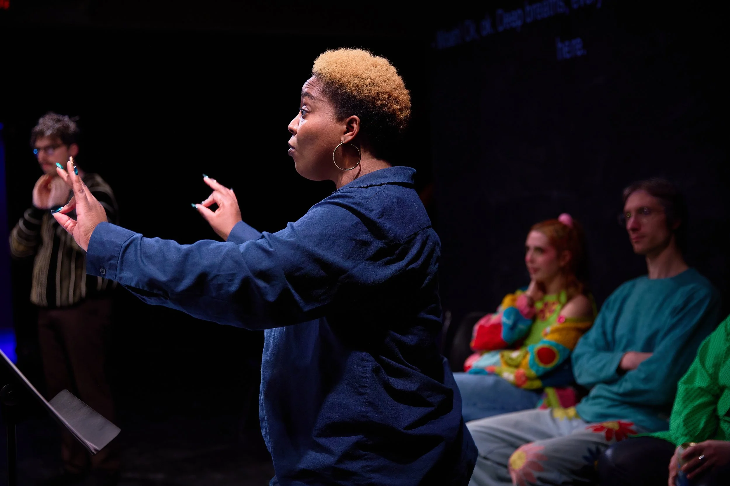 Une femme noire en train de diriger une activité avec plusieurs personnes assises en arrière-plan, dans un environnement sombre.