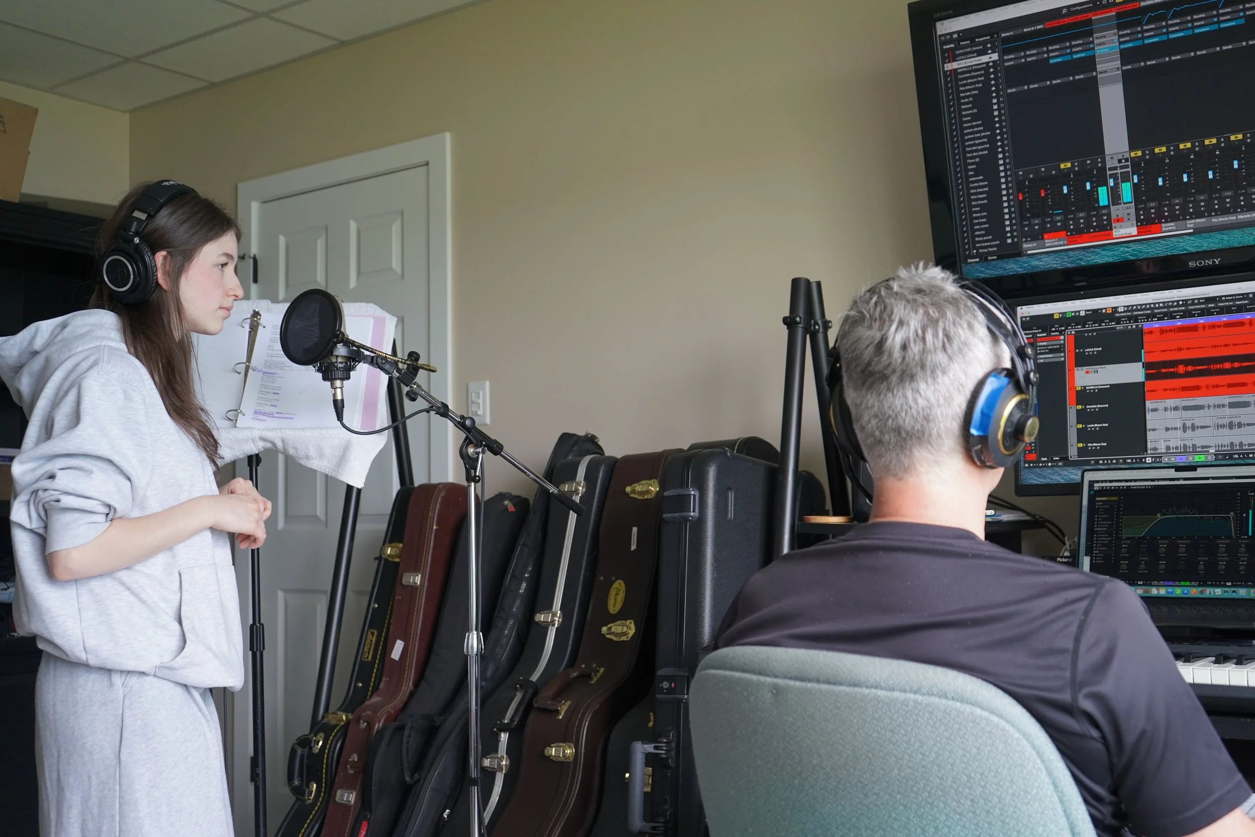 Deux personnes portent des écouteurs et travaillent dans un studio de musique avec des ordinateurs affichant des logiciels de production musicale, une personne chante dans un micro, une autre joue du piano.