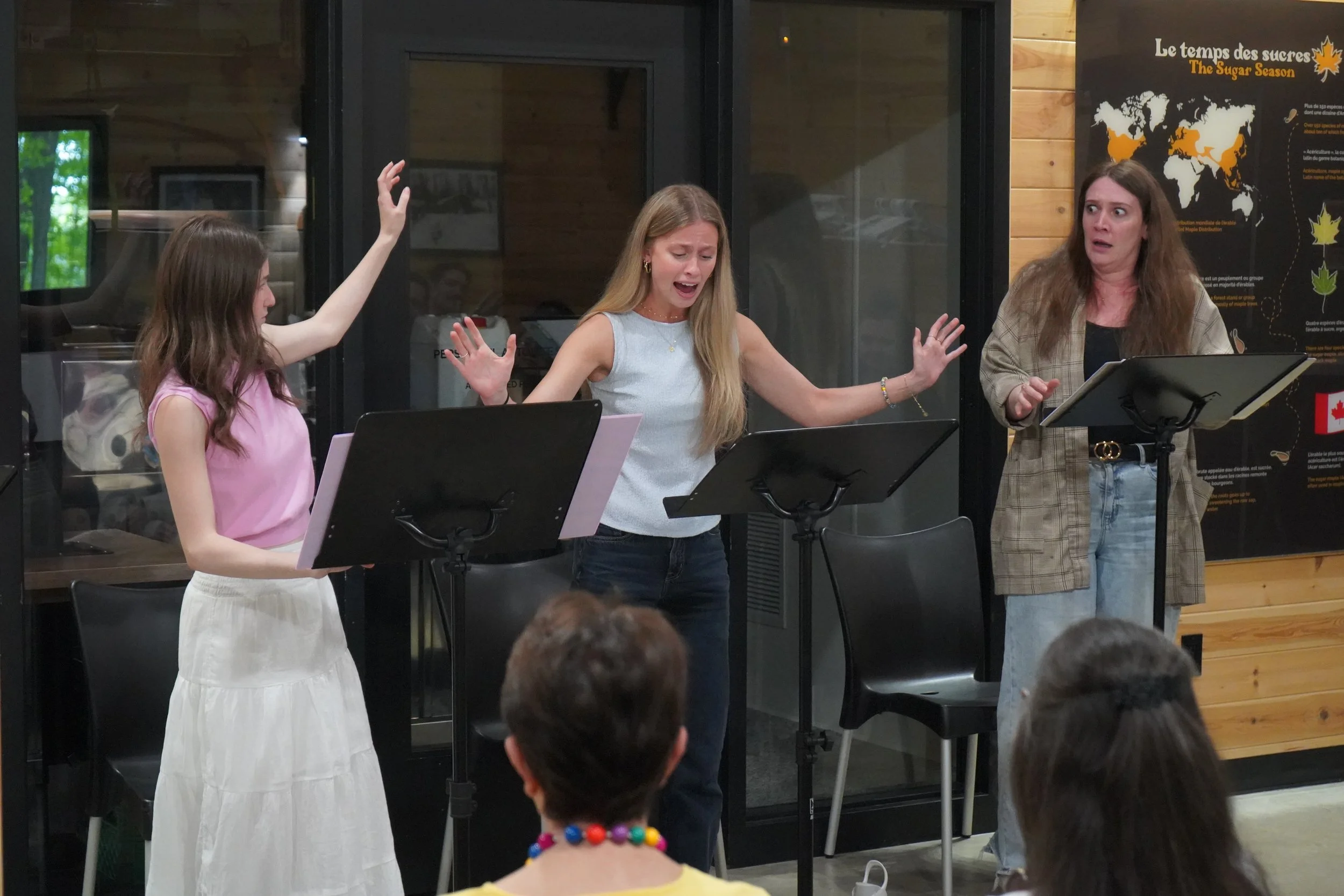 Trois femmes en train de chanter ou de réciter, avec un public devant elles. Derrière elles, un panneau noir avec une carte du monde et du texte en français et en anglais sur la saison sucrière.