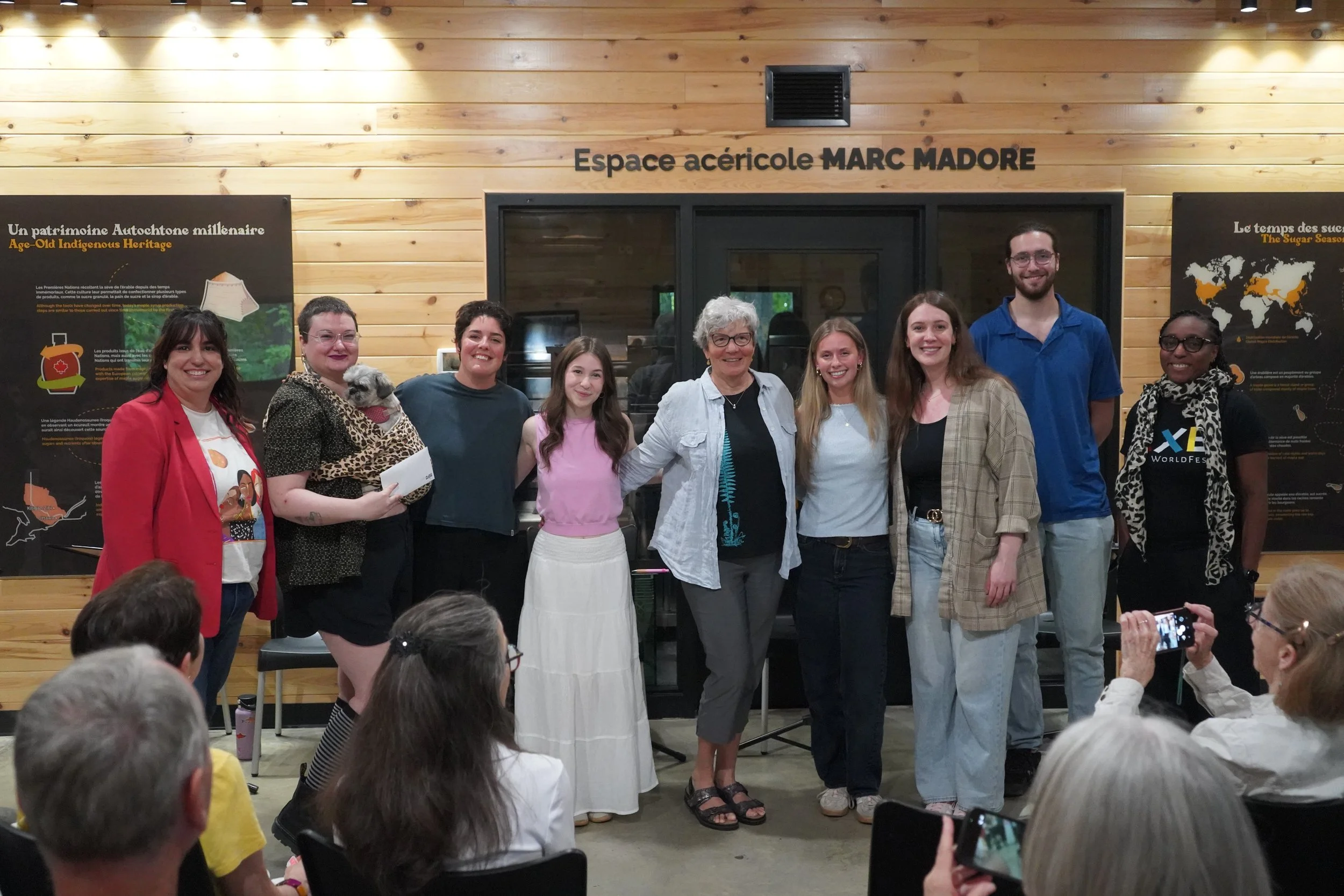 Groupe de neuf personnes posant ensemble à l'intérieur d'une salle en bois, devant un mur avec panneaux d'informations, quelques participants regardent la scène et d'autres prennent des photos.