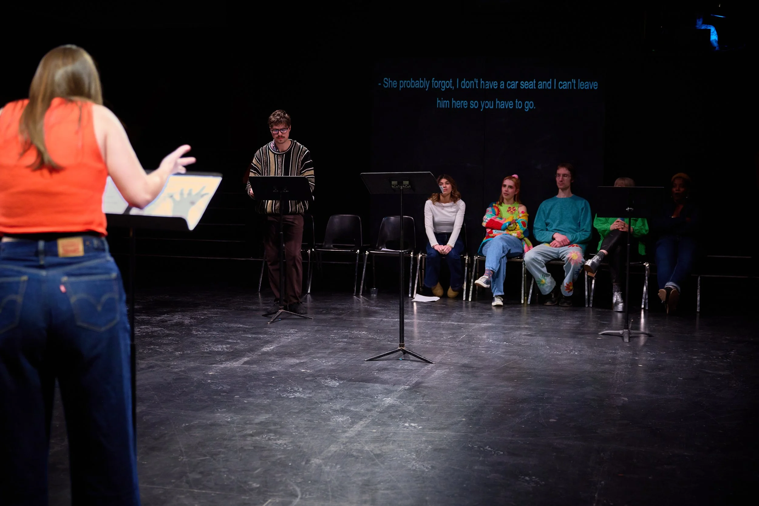 Une scène de théâtre avec une femme en premier plan parlant et six personnes assises en arrière-plan, certains en couleurs vives, avec des sous-titres en anglais projetés derrière eux.