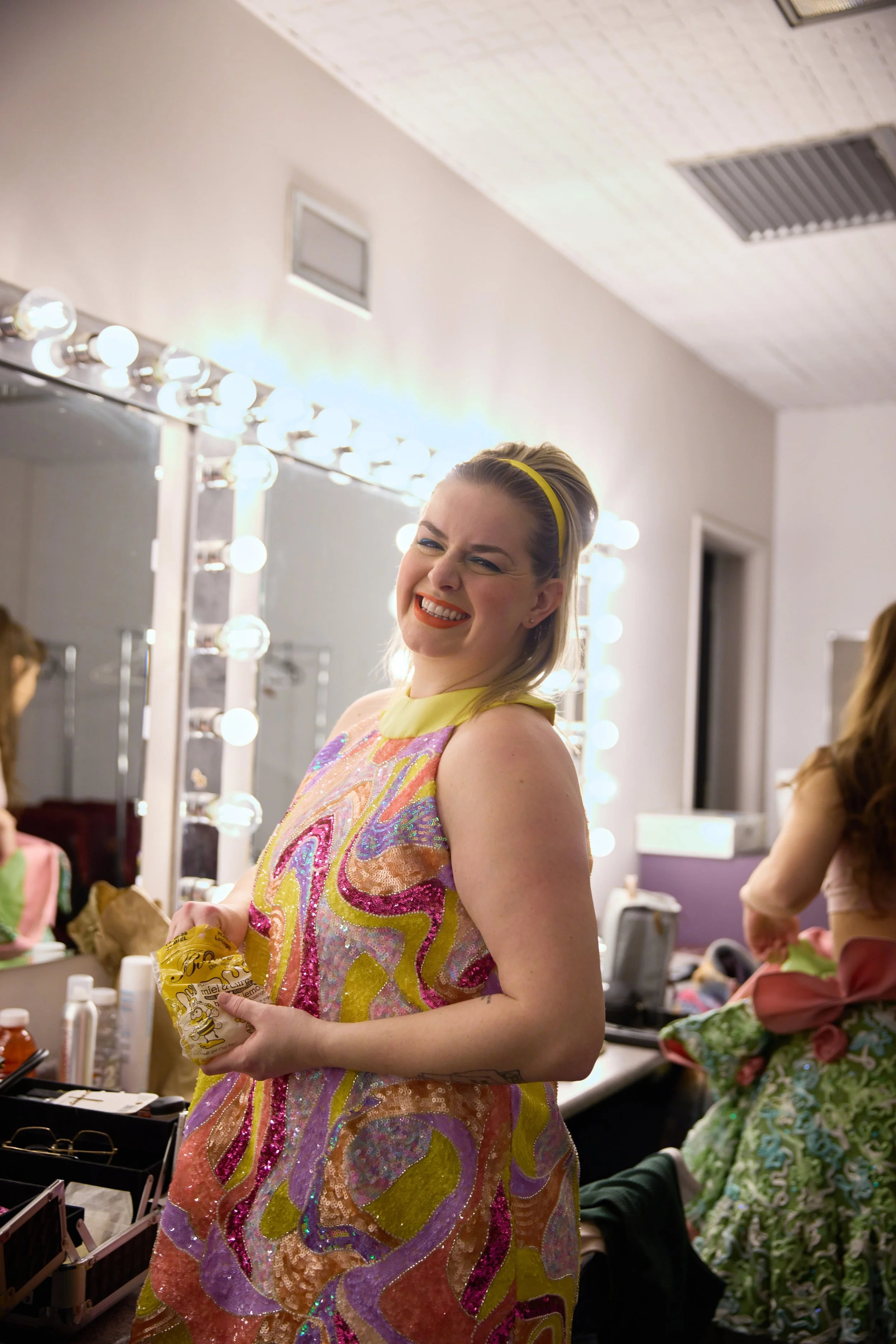 Une femme souriante en robe colorée dans une loge de maternité avec un miroir éclairé.