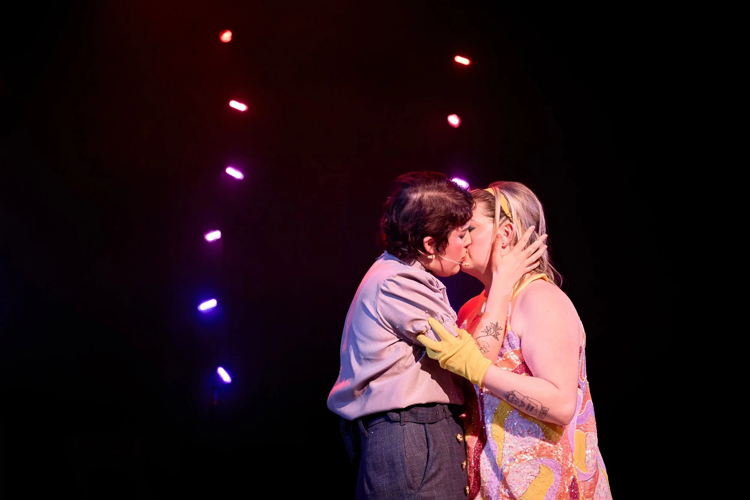 Deux femmes s'embrassent sur scène, éclairage coloré en arrière-plan.