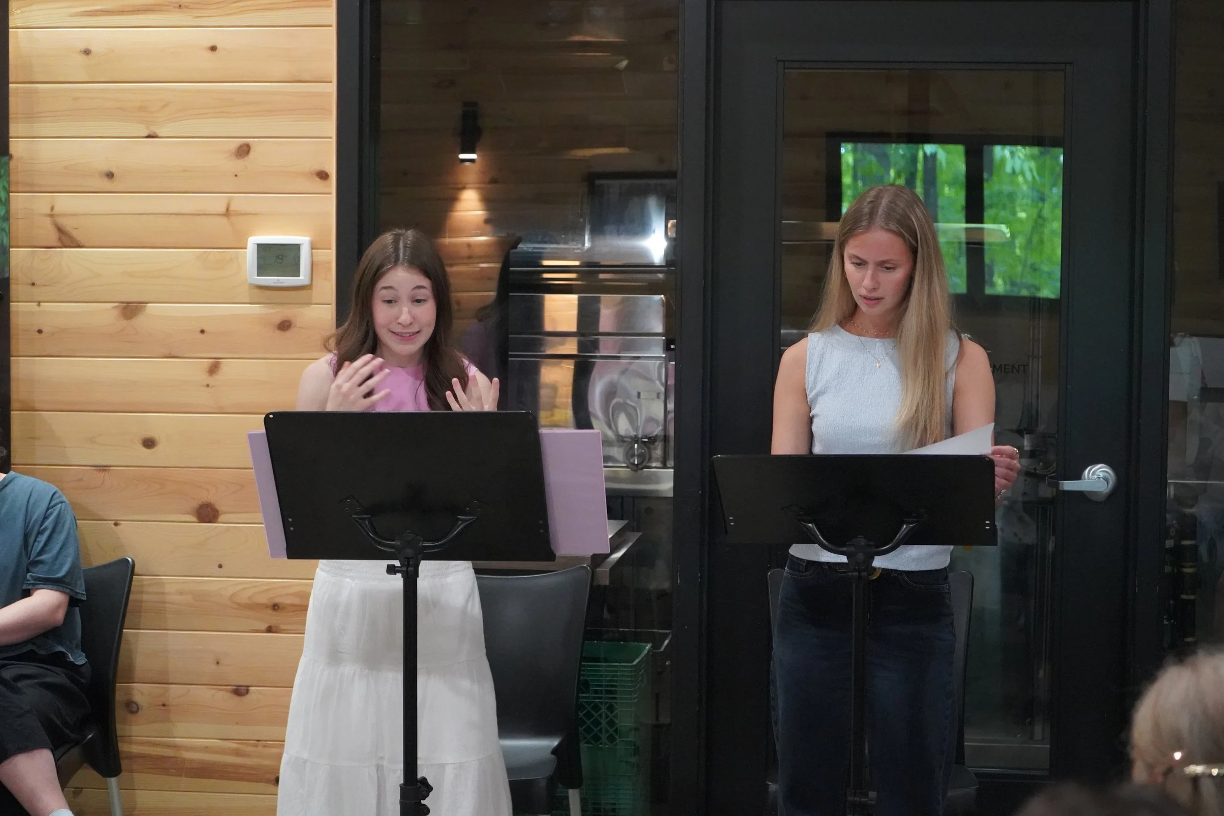 Deux jeunes femmes se tiennent à côté de partitions de musique sur des supports à trépied, dans une pièce avec des murs en bois et des fenêtres.