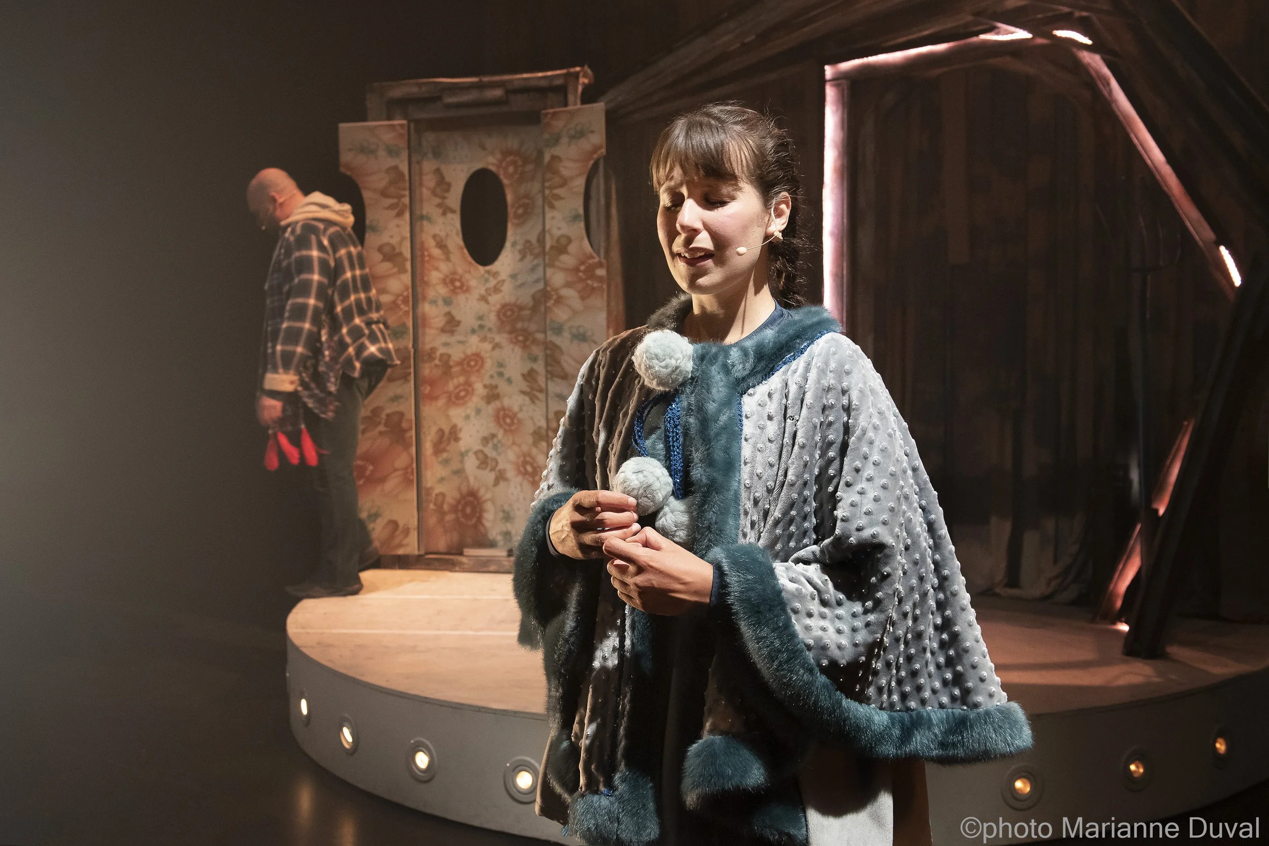 Une jeune femme avec une veste en fourrure grise et bleue, se tenant sur scène avec un micro attaché au visage, semblant émue, en train de jouer une scène théâtrale. En arrière-plan, un homme plus âgé portant une veste à carreaux et une écharpe rouge