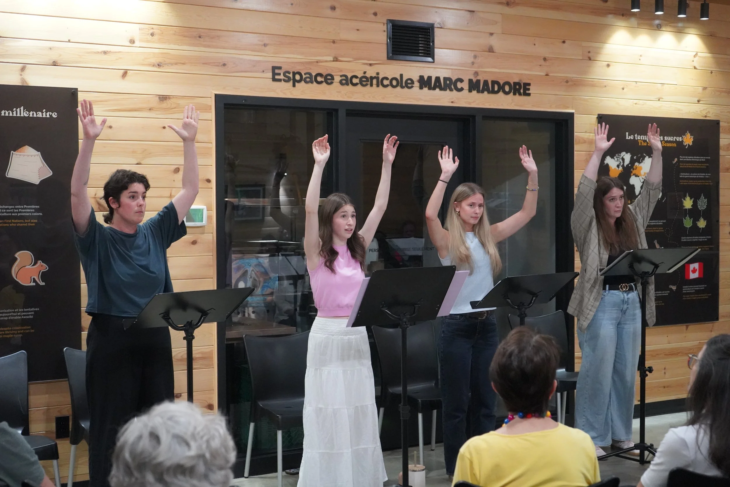 Quatre jeunes femmes se tiennent derrière des pupitres, levant une main pour répondre ou participer, dans une salle avec un mur en bois et un signe indiquant 'Espace acéricole MARC MADORE'. Des personnes sont assises dans l'audience, regardant la scè