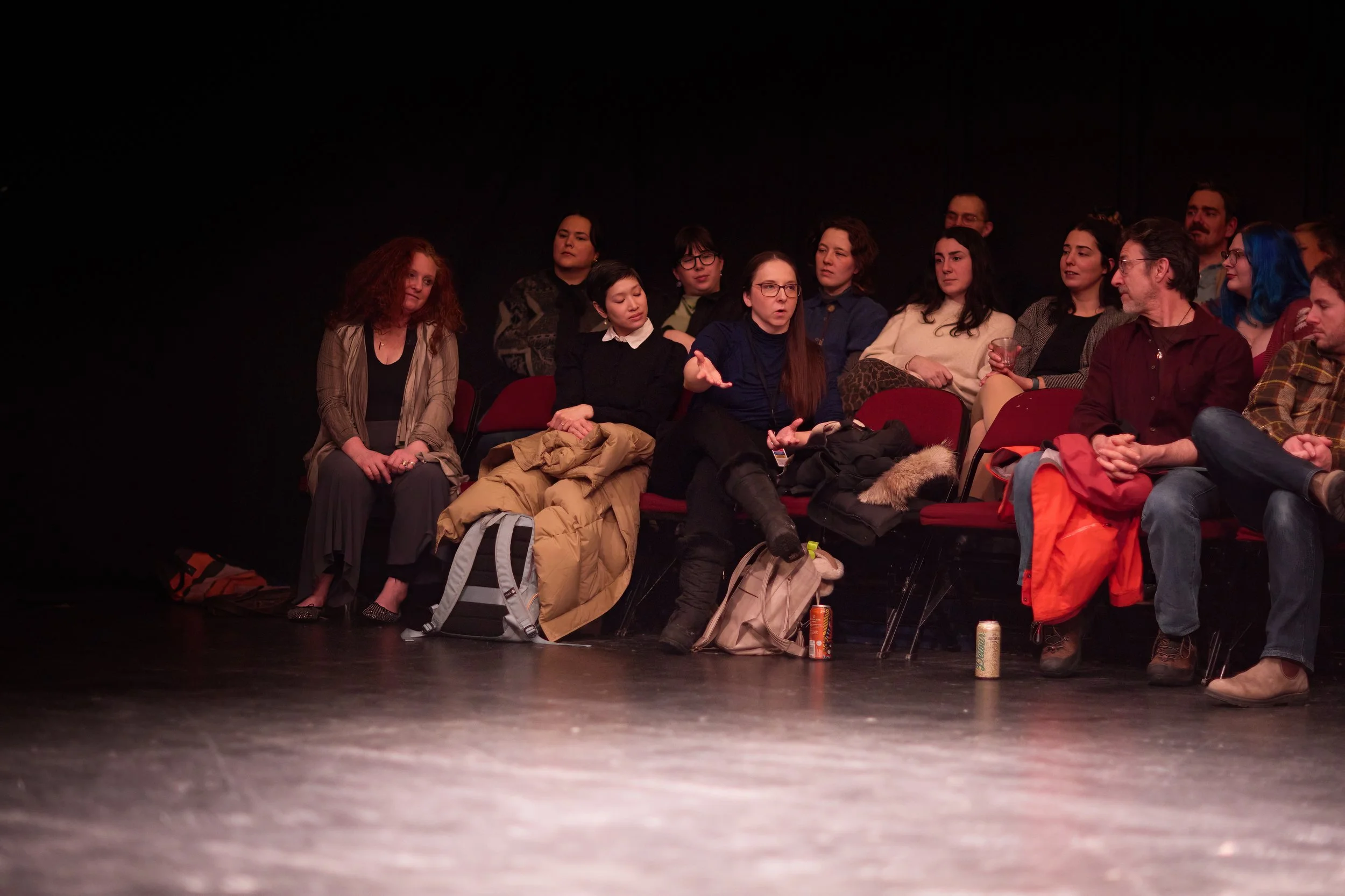 Groupe de personnes assises dans une salle sombre, discutant ou écoutant une présentation, certaines avec des sacs à leurs pieds.