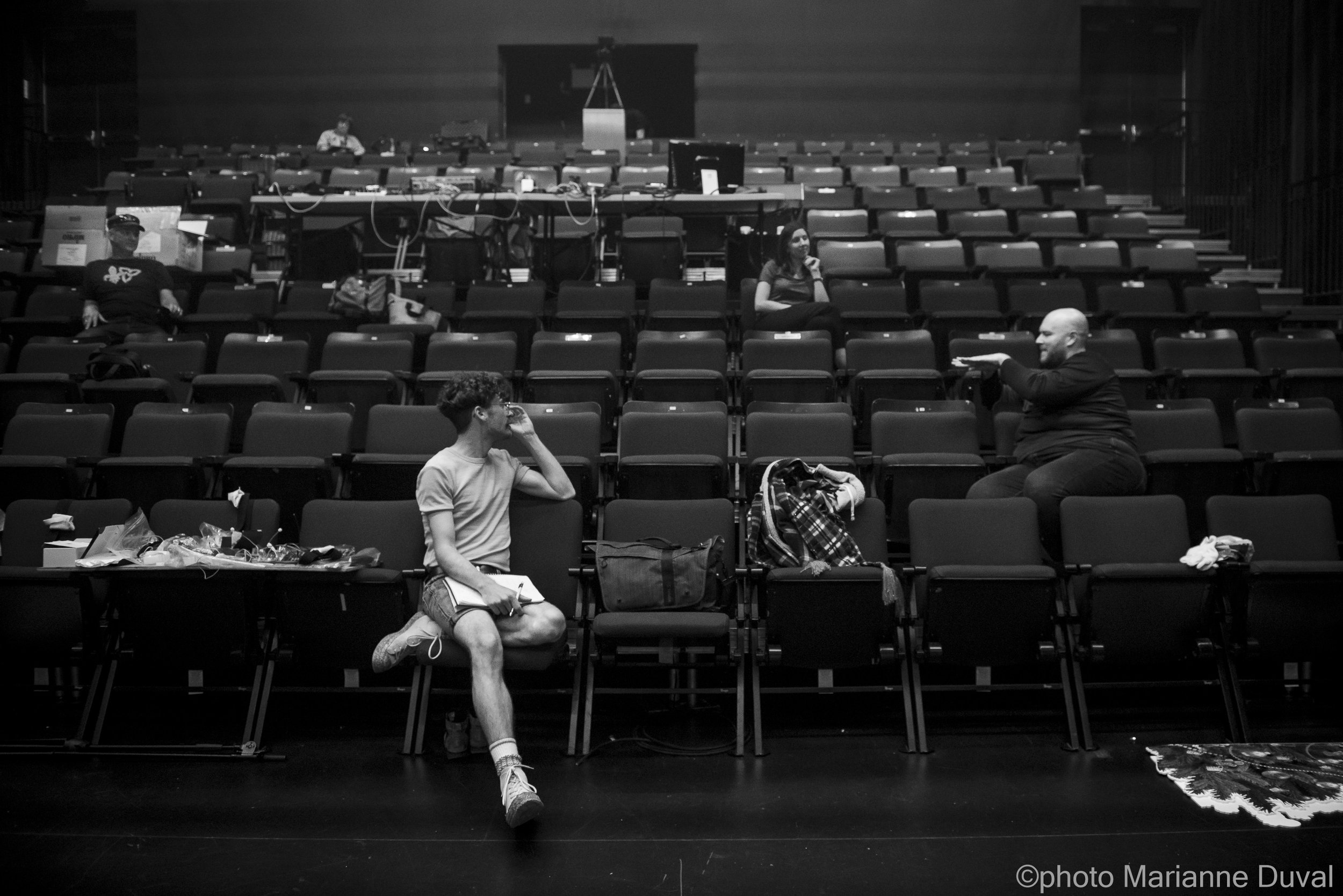 Une salle de conférence ou de théâtre avec quelques personnes assises, discutant ou préparant des présentations, y compris un jeune homme en premier plan, une femme en haut, et un homme qui parle ou donne des instructions.