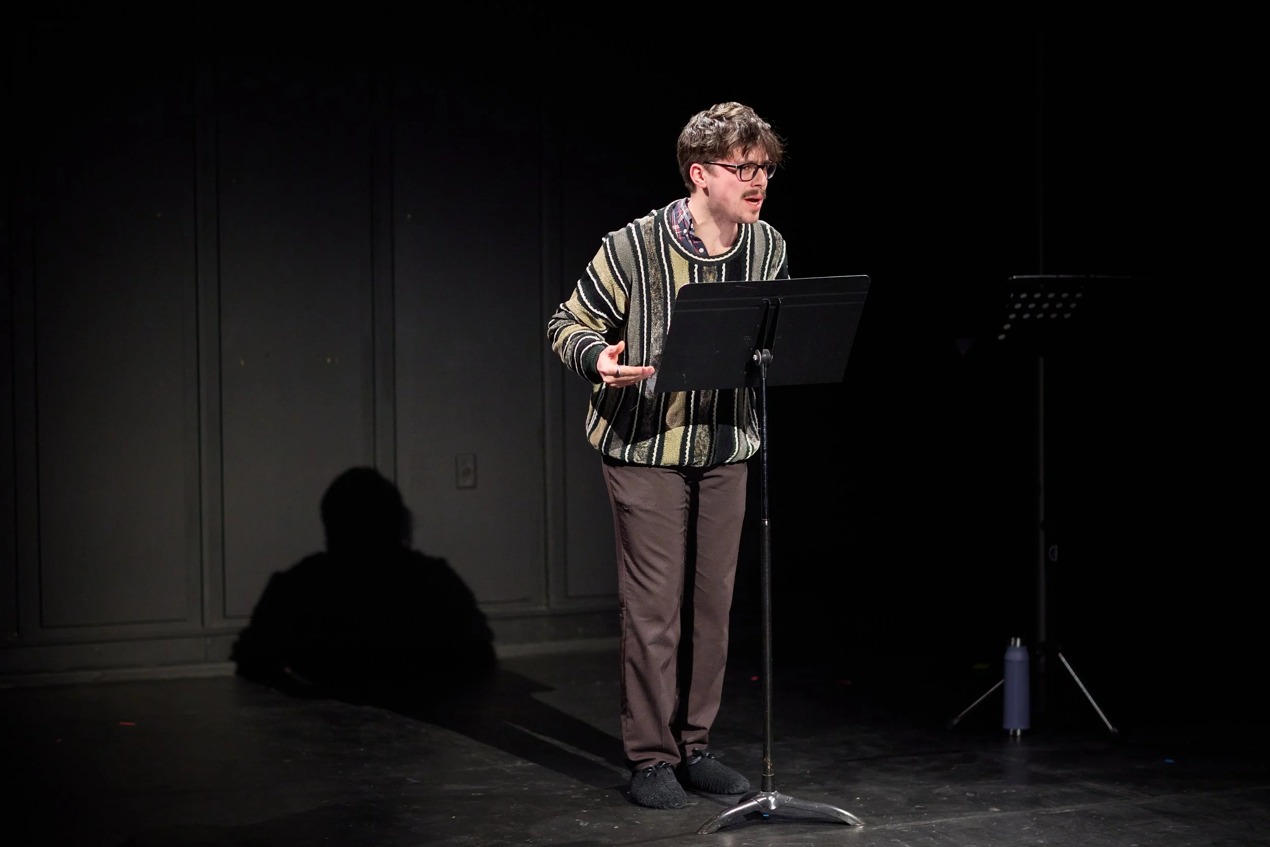 Un homme parlant devant un pupitre avec une partition, sur scène en studio noir.