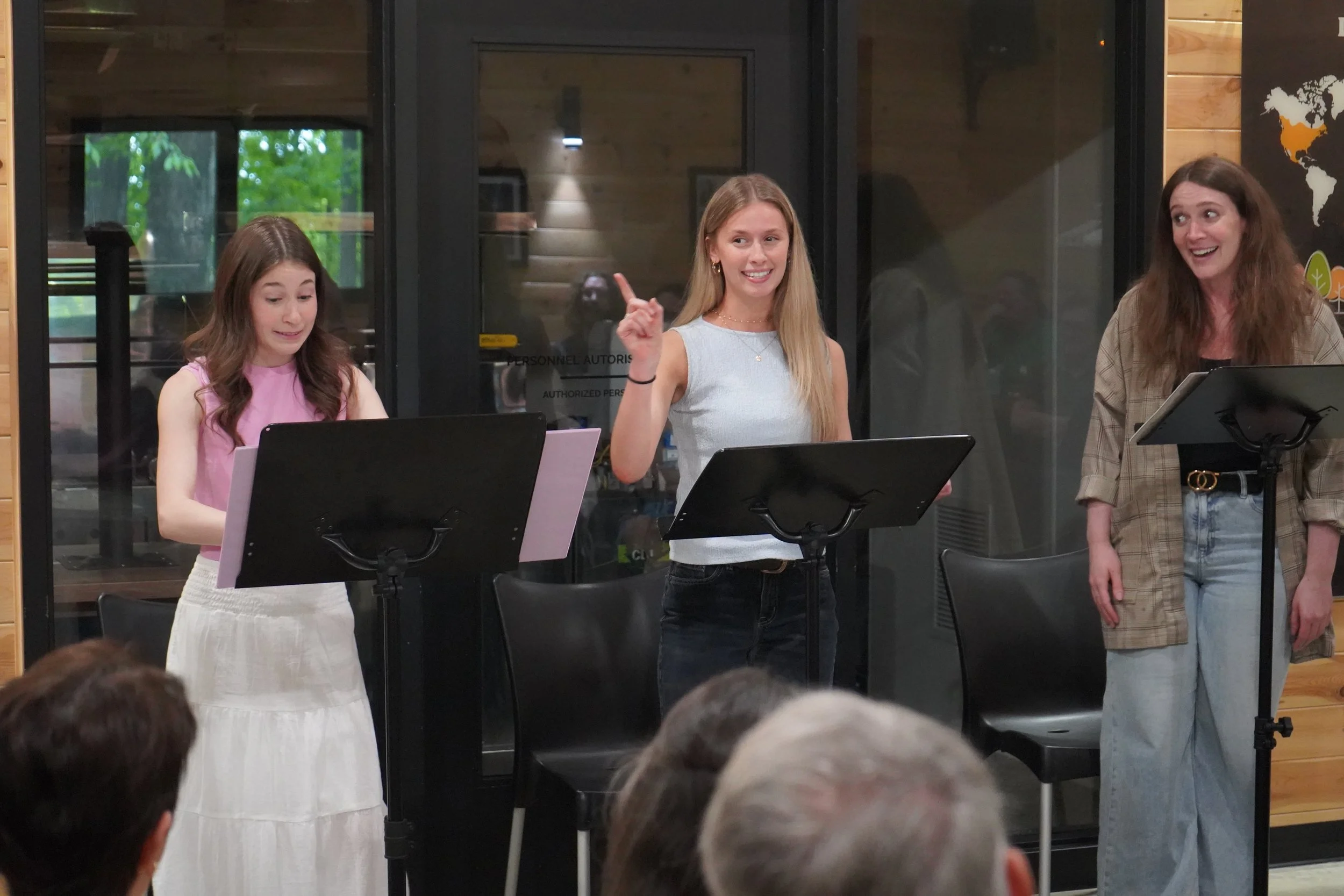 Trois femmes se tiennent devant un pupitre à musique, parlant ou chantant, dans une pièce avec un mur en bois et des écrans en verre, et une carte du monde décorative.