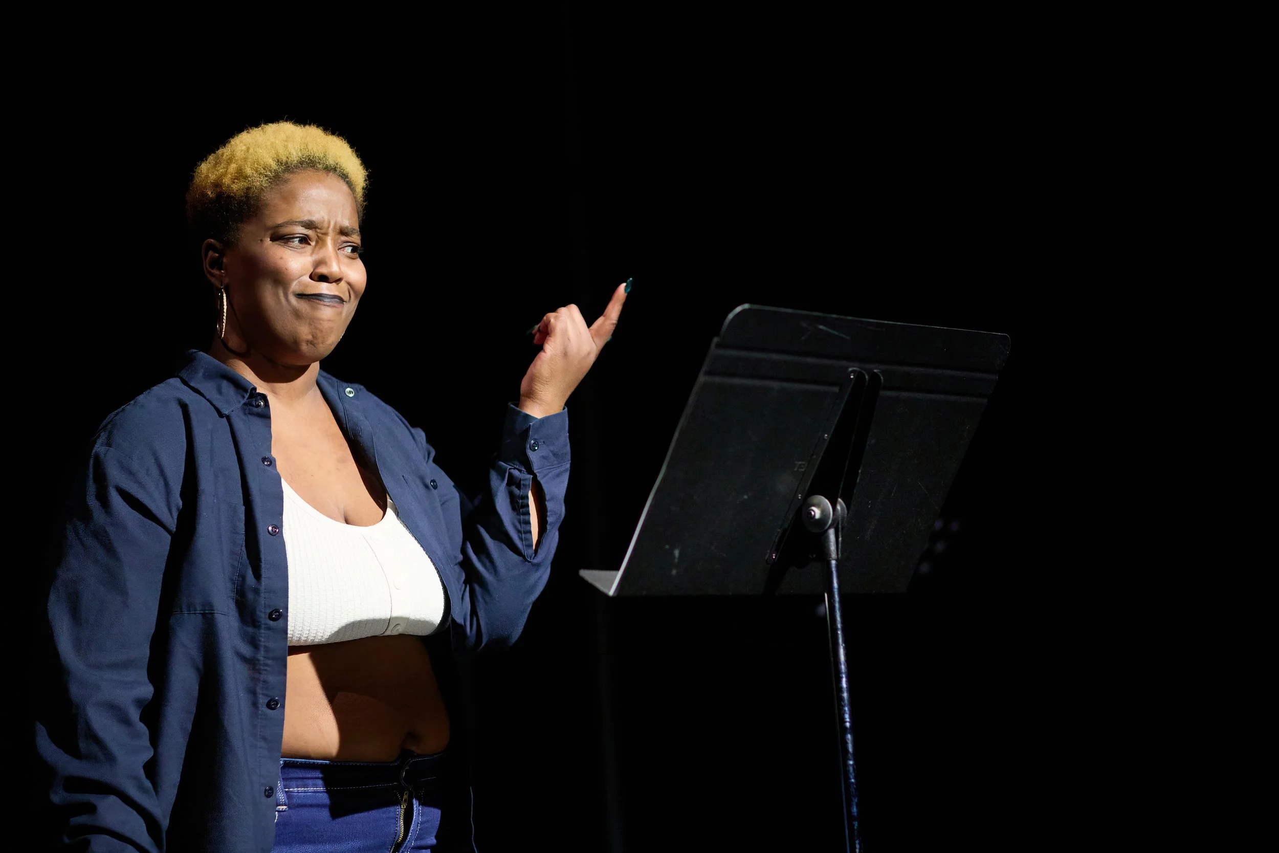 Une femme avec des cheveux blonds courts, portant un chemisier blanc et une veste bleue, semble faire une expression confuse ou sceptique lors d'une lecture ou performance, avec un pupitre noir devant elle sur un fond sombre.