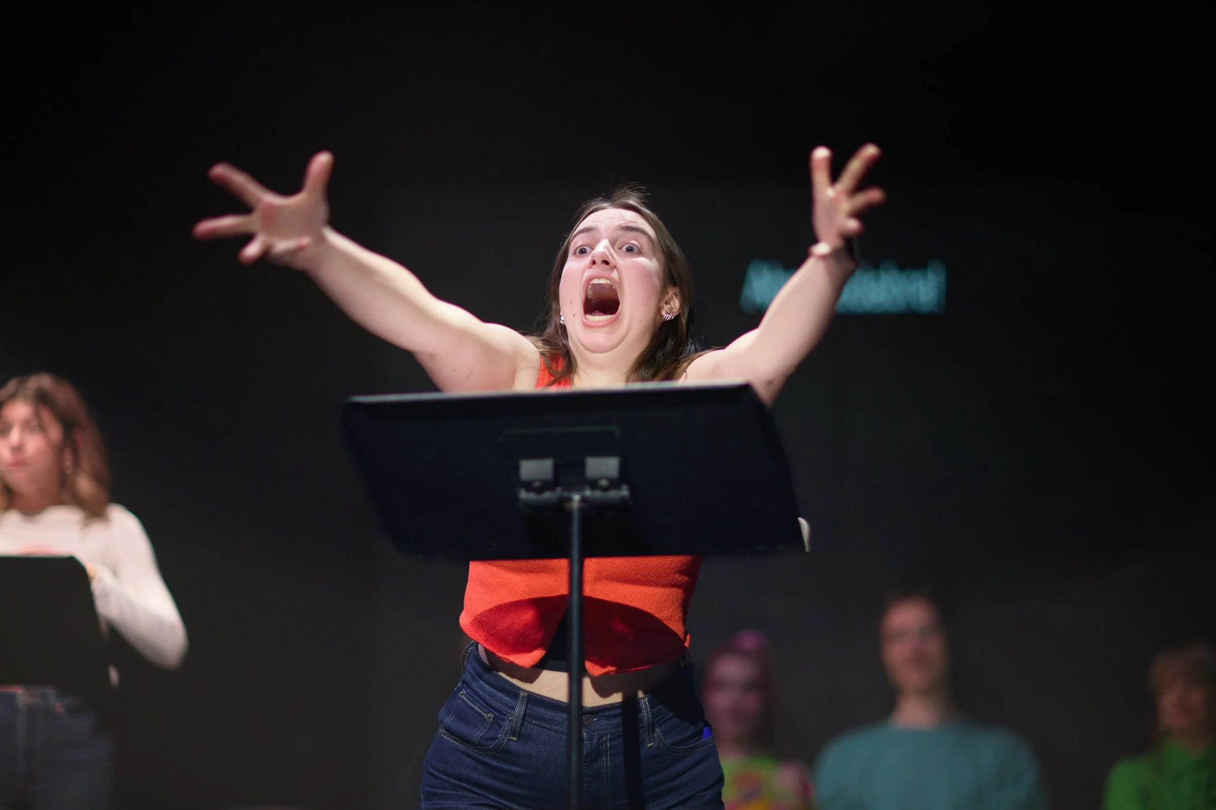 Une jeune femme parle ou chante avec une expression émotionnelle forte, les bras levés, devant un pupitre, avec d'autres personnes en arrière-plan.
