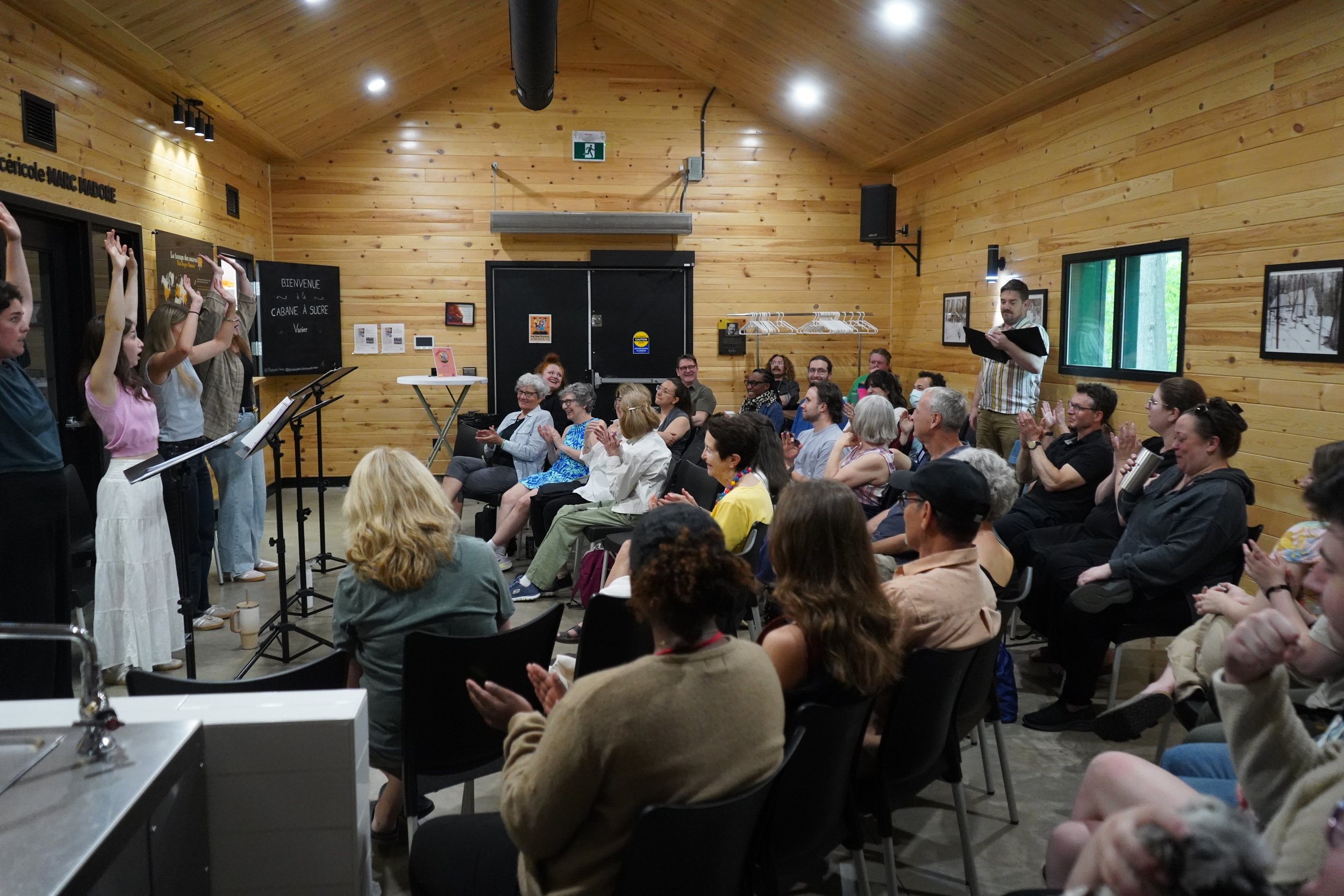 Une salle avec un public assis, applaudissant, où se déroule un événement ou une présentation ; à l'avant, plusieurs jeunes femmes sont debout, s'adressant à l'audience. Les murs sont en bois, avec des fenêtres et quelques affiches ou images.