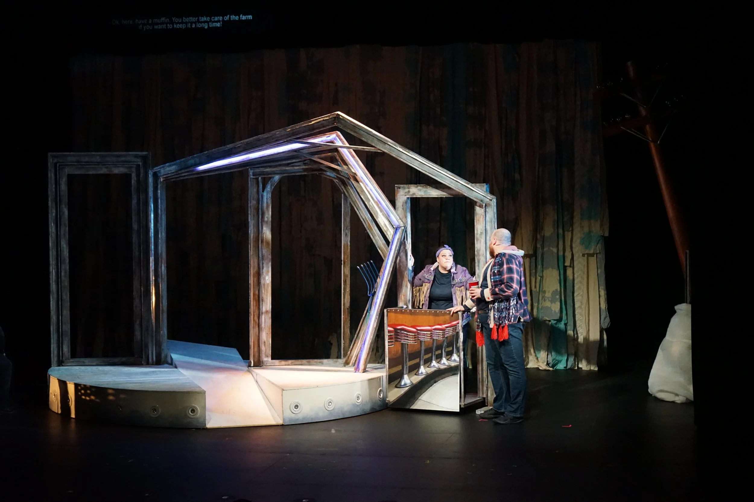 Deux personnes discutent sur une scène de théâtre avec une structure en bois. Un des deux porte un manteau à carreaux. La scène est décorée de rideaux et d'autres éléments de décor.