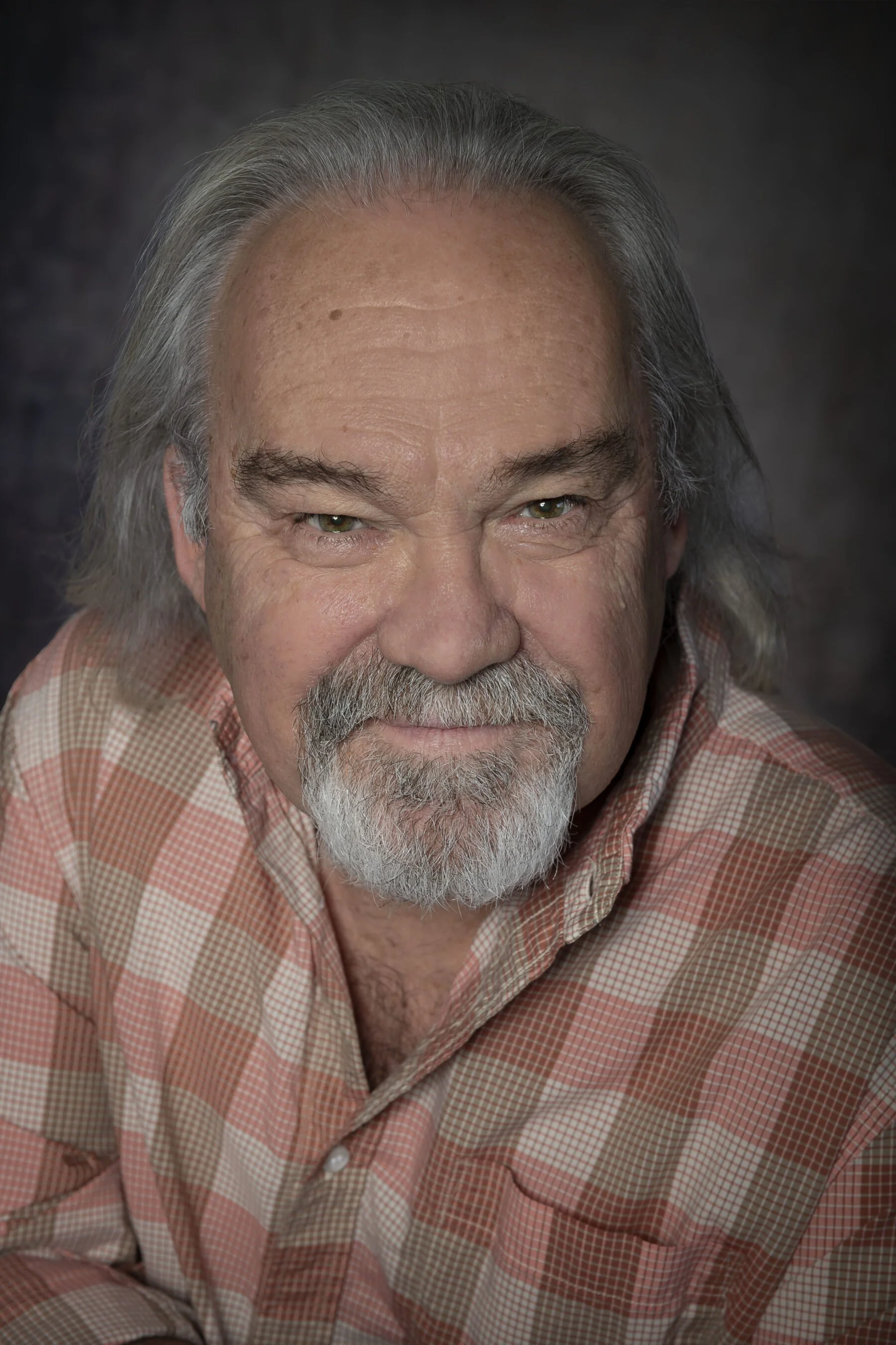 portrait d'un homme âgé avec des cheveux gris, une barbe grise, portant une chemise à carreaux rose et beige, sourire léger.