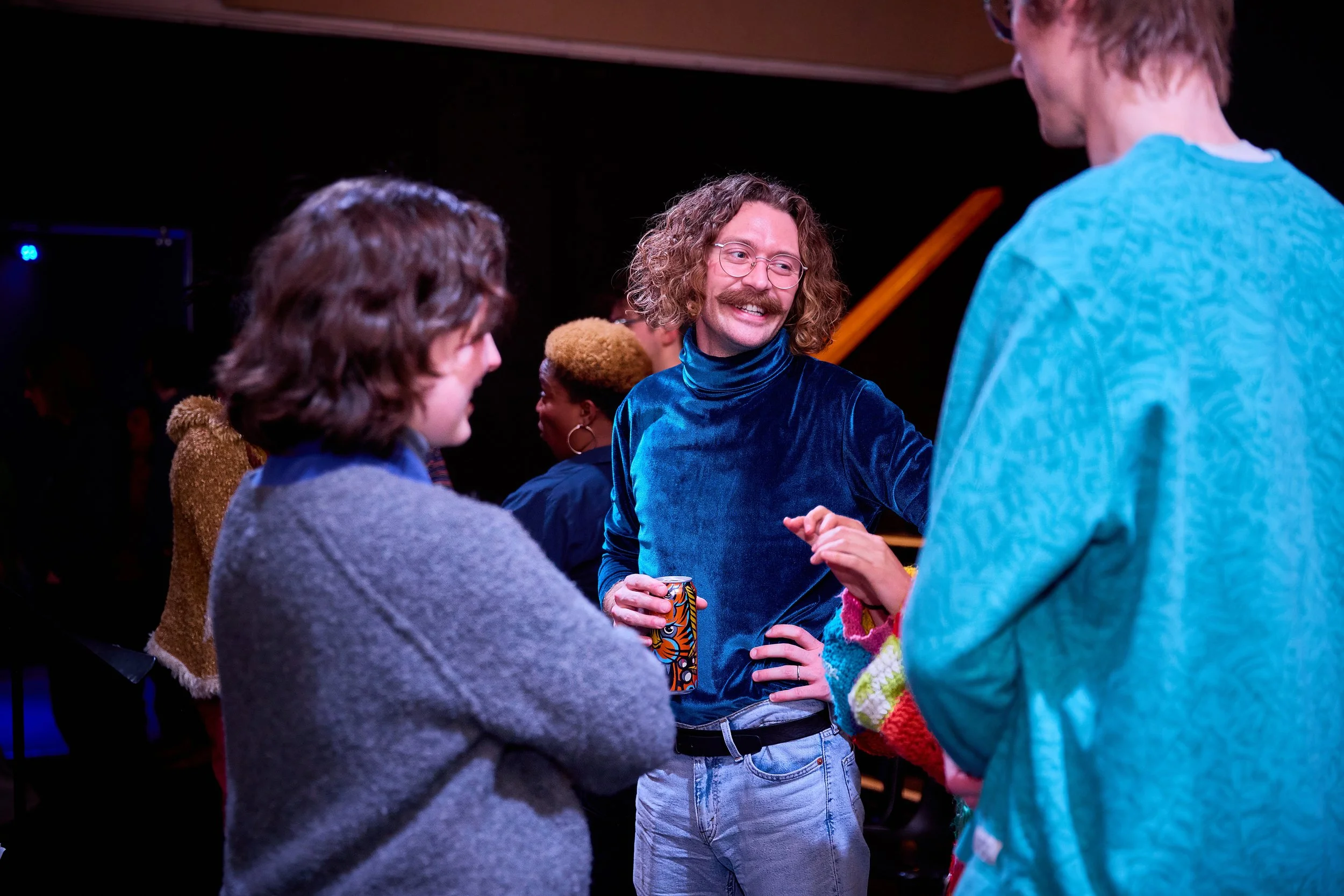 Groupe de jeunes personnes discutant dans une pièce sombre, ambiance décontractée, avec une personne portant un haut en velours bleu et tenant une canette.