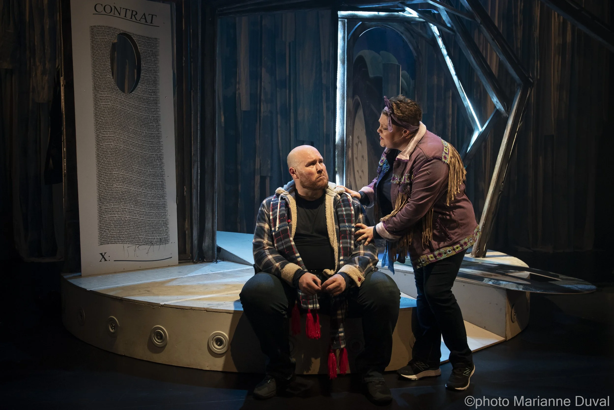 Deux acteurs sur scène, un homme assis et une femme debout, en pleine représentation théâtrale. L'homme porte une veste à carreaux et un hoodie, la femme porte une veste en velours avec des franges, et elles semblent engager une scène intense.