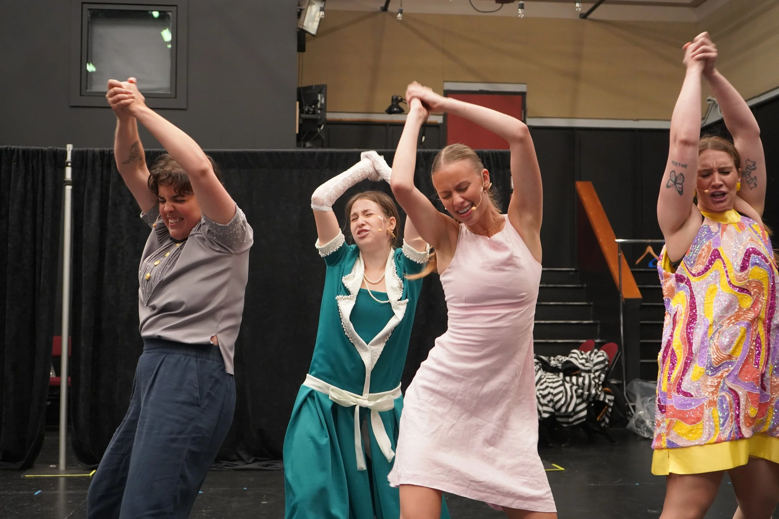 Quatre femmes en vêtements de spectacle ou de costume, tenant leurs mains levées et semblant chanter ou danser sur scène dans un théâtre ou une salle de spectacle.