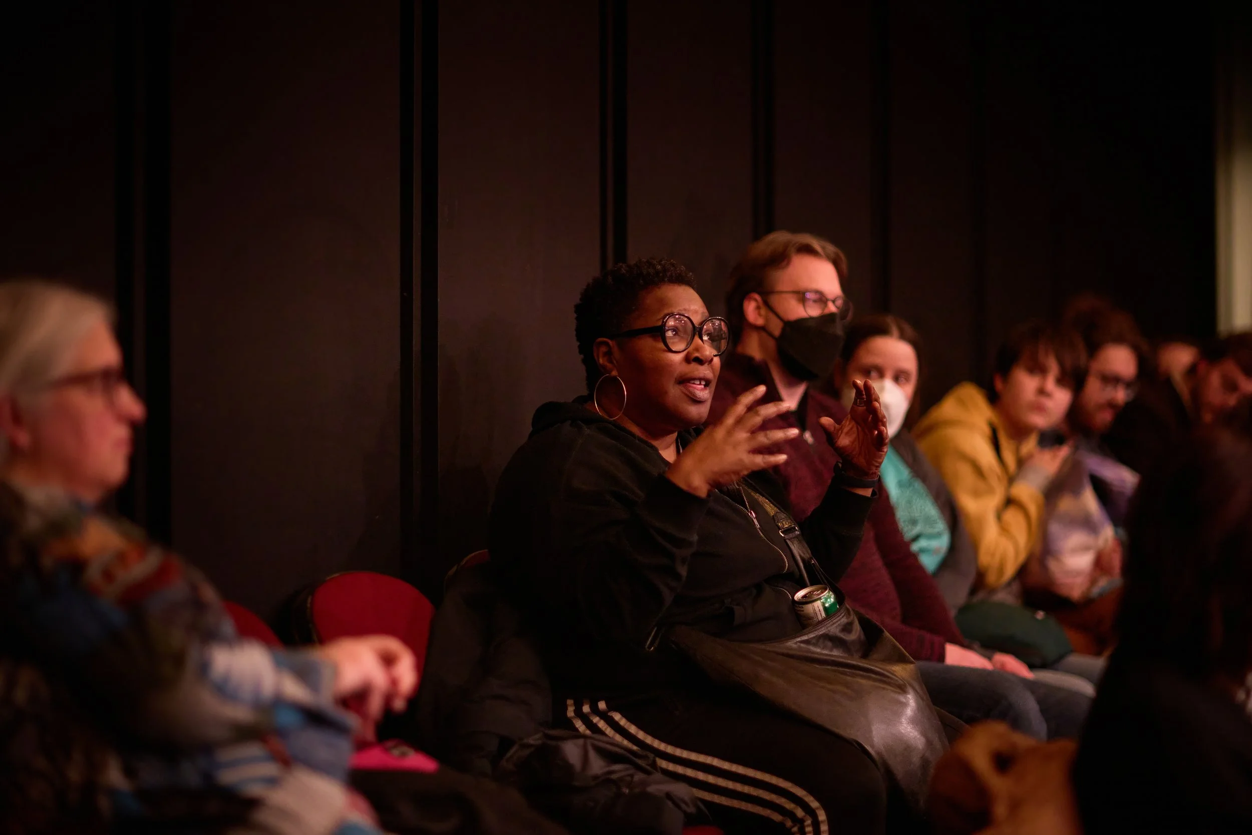 Groupe de personnes assistant à une discussion ou un événement, avec une femme noire en avant qui parle et utilise des gestes, portant des lunettes, vêtue de noir, entourée de personnes portant des masques.