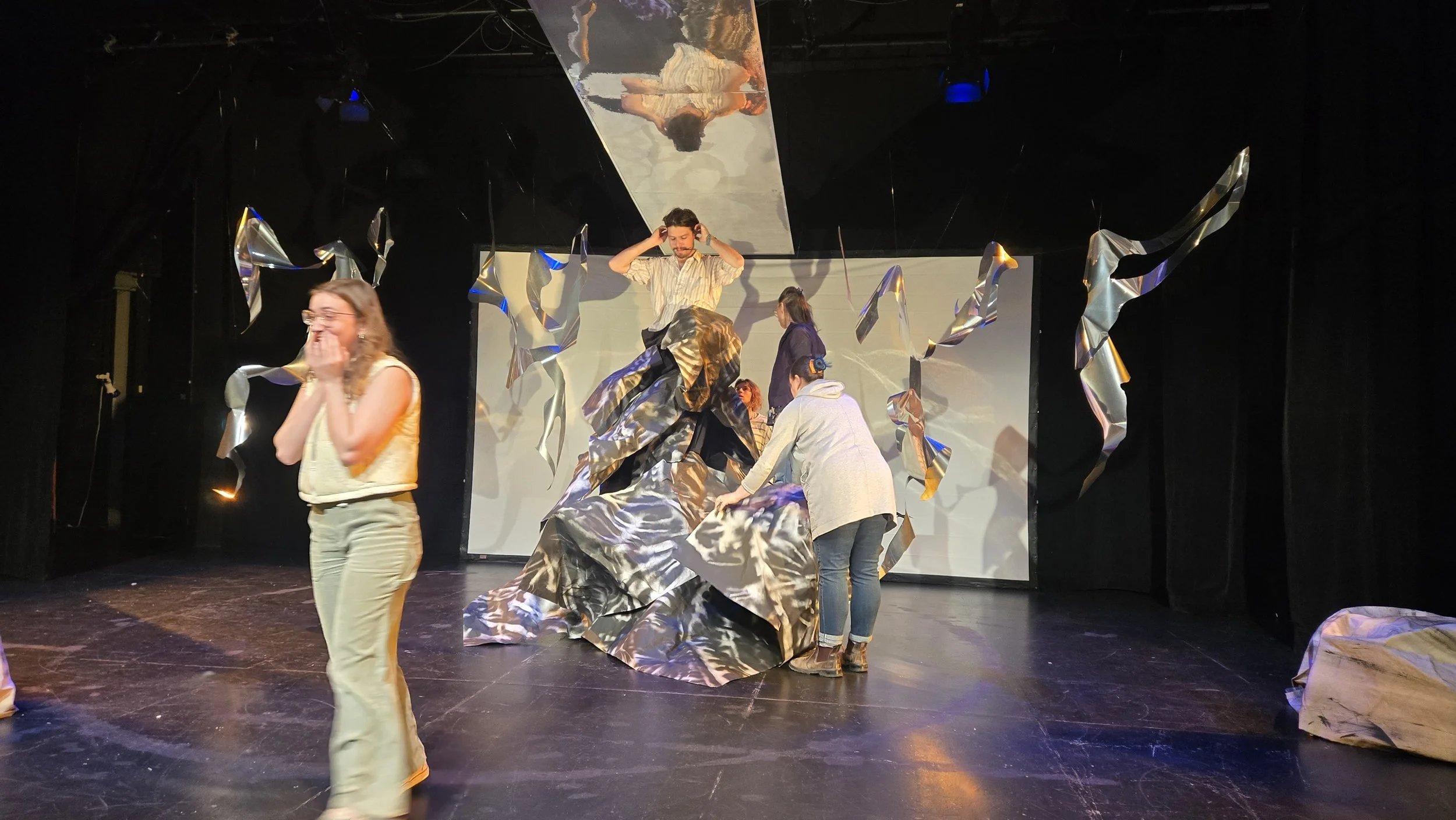 Marée haute. Scène de théâtre avec plusieurs personnes, quelques-unes en train de préparer une scène, décor en papier ou tissu avec des éléments argentés suspendus et une personne en costume sur une plateforme.