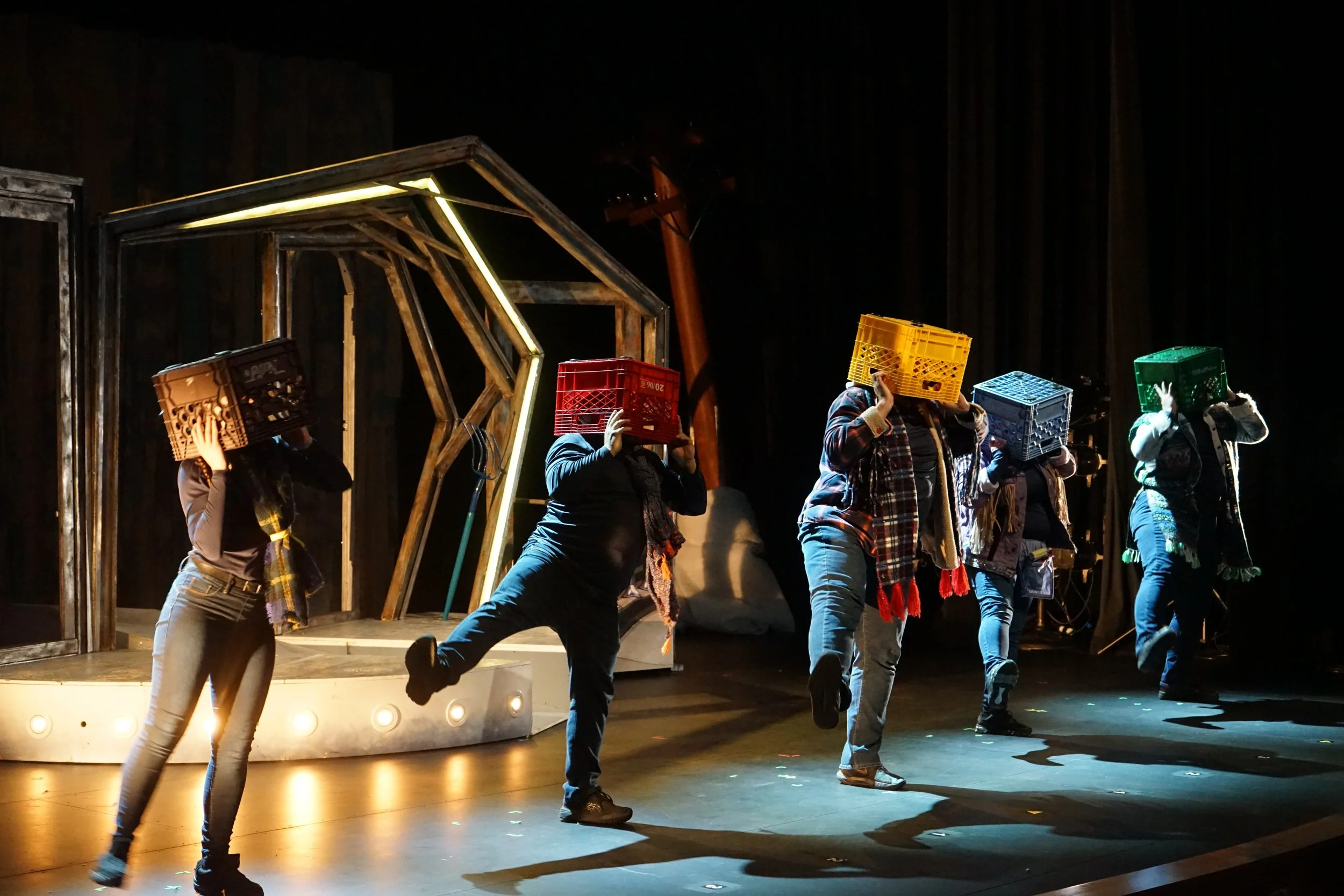Des personnes sur scène portant des caisses en plastique sur la tête, dans un décor de théâtre avec des éléments en bois et un éclairage dramatique.