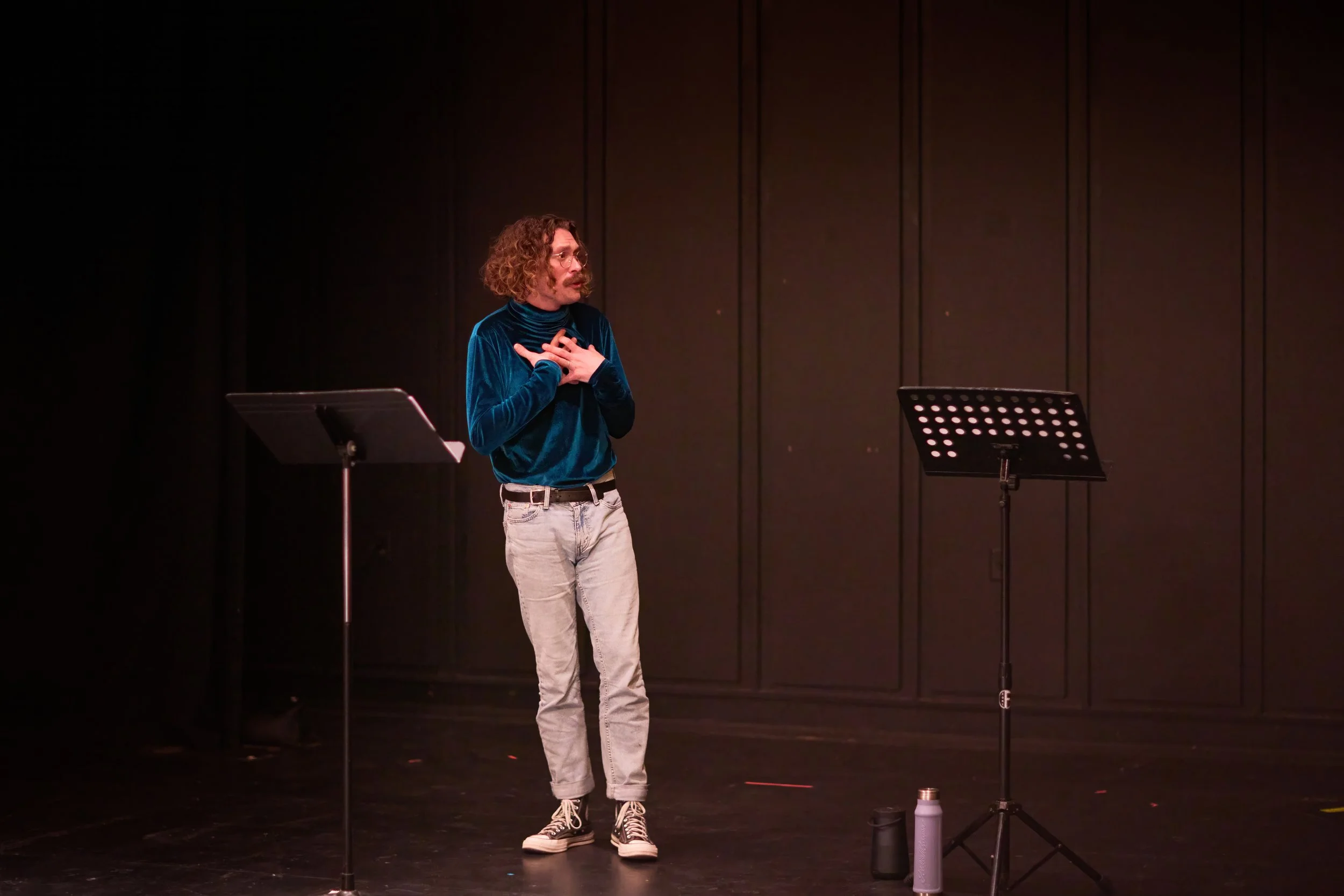 Un homme avec des cheveux bouclés et une moustache, portant un pull en velours bleu, des jeans gris et des baskets noires et blanches, se tient sur scène avec un micro, deux supports de partition vides, une bouteille thermos et une gourde à côté, dan