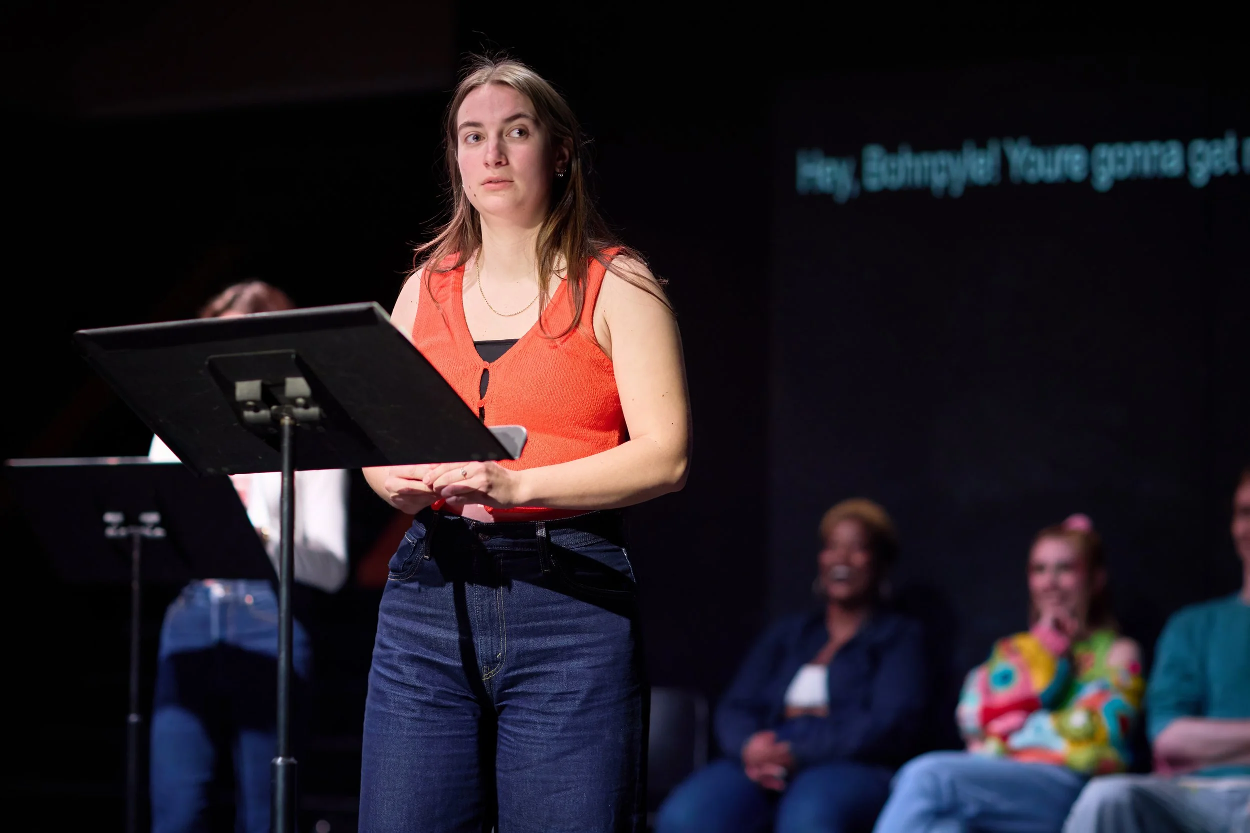 Une jeune femme en haut d'une scène, tenant un pupitre, dans une salle sombre avec un public assis en arrière-plan, regardant vers elle. Un écran affiche un texte en anglais.